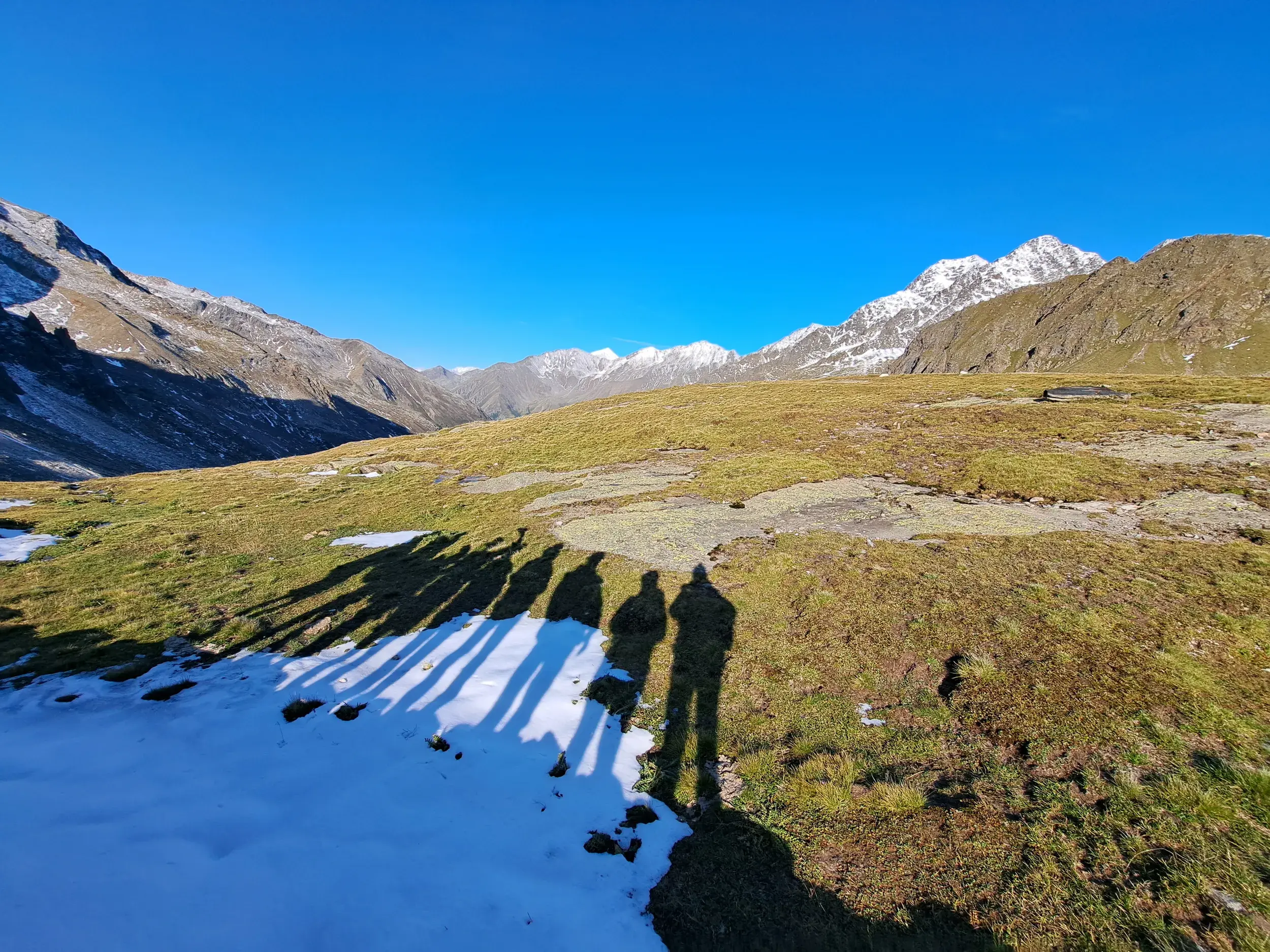 Schatten einer Wandergruppe auf einer Almwiese mit Schnee vor der Bergkulisse der Texelgruppe in Südtirol.