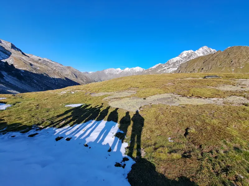 Schatten einer Wandergruppe auf einer Almwiese mit Schnee vor der Bergkulisse der Texelgruppe in Südtirol.
