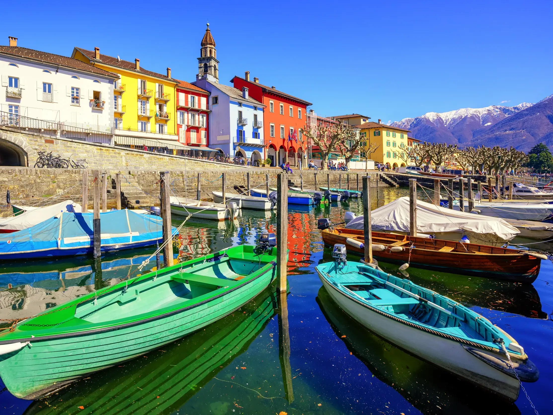 Farbenfrohe Boote im Hafen von Ascona spiegeln sich im Wasser des Lago Maggiore vor einer bunten Häuserzeile und Alpenpanorama.