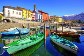 Farbenfrohe Boote im Hafen von Ascona spiegeln sich im Wasser des Lago Maggiore vor einer bunten Häuserzeile und Alpenpanorama.