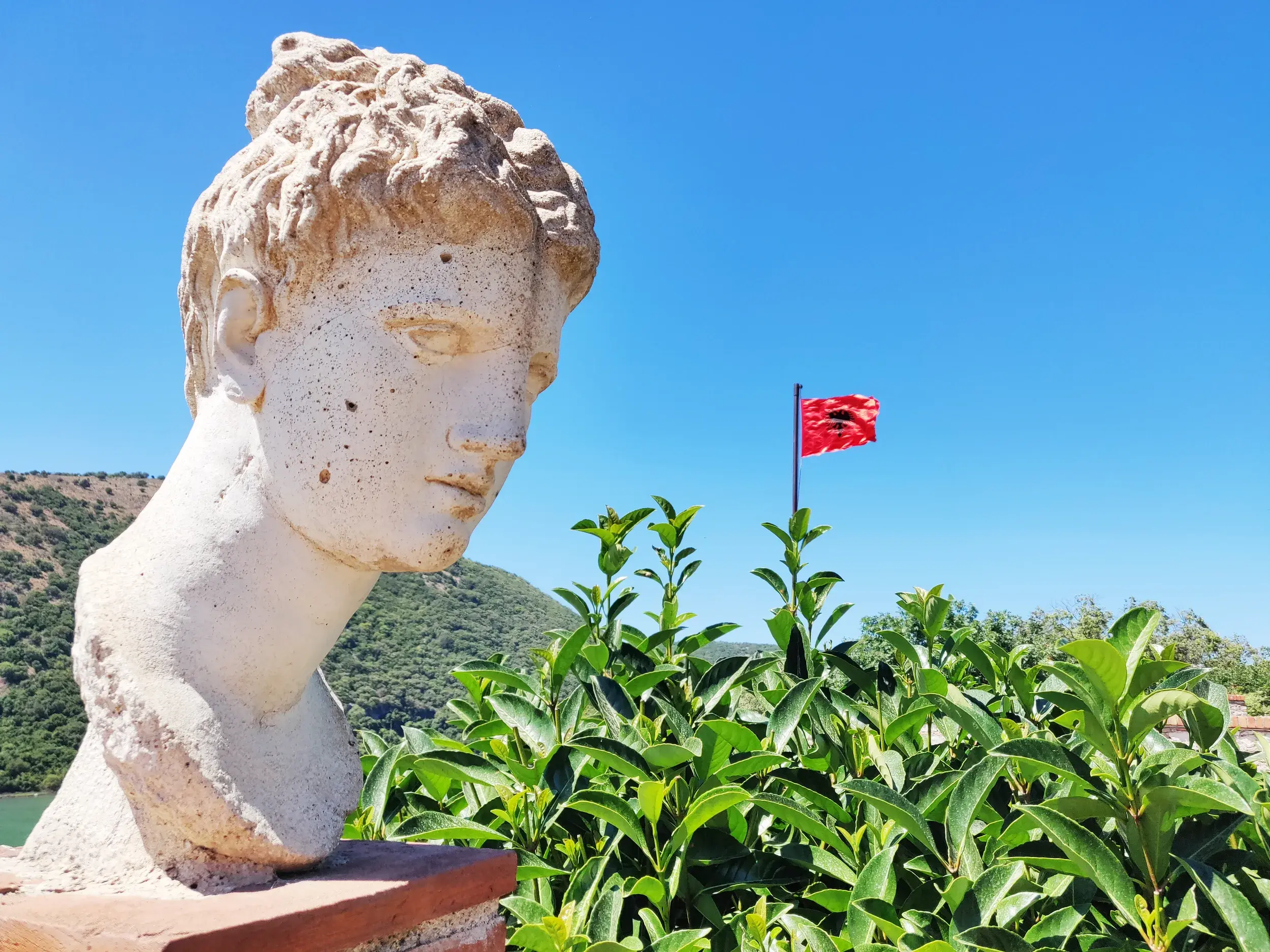 Antike Steinbüste in der Ruinenstadt Butrint in Albanien, im Hintergrund weht die albanische Flagge vor blauem Himmel.
