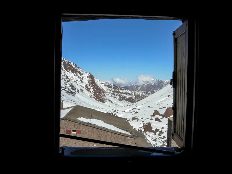 Blick aus dem Fenster einer Berghütte auf die sonnige, schneebedeckte Gipfellandschaft des Hohen Atlas in Marokko.