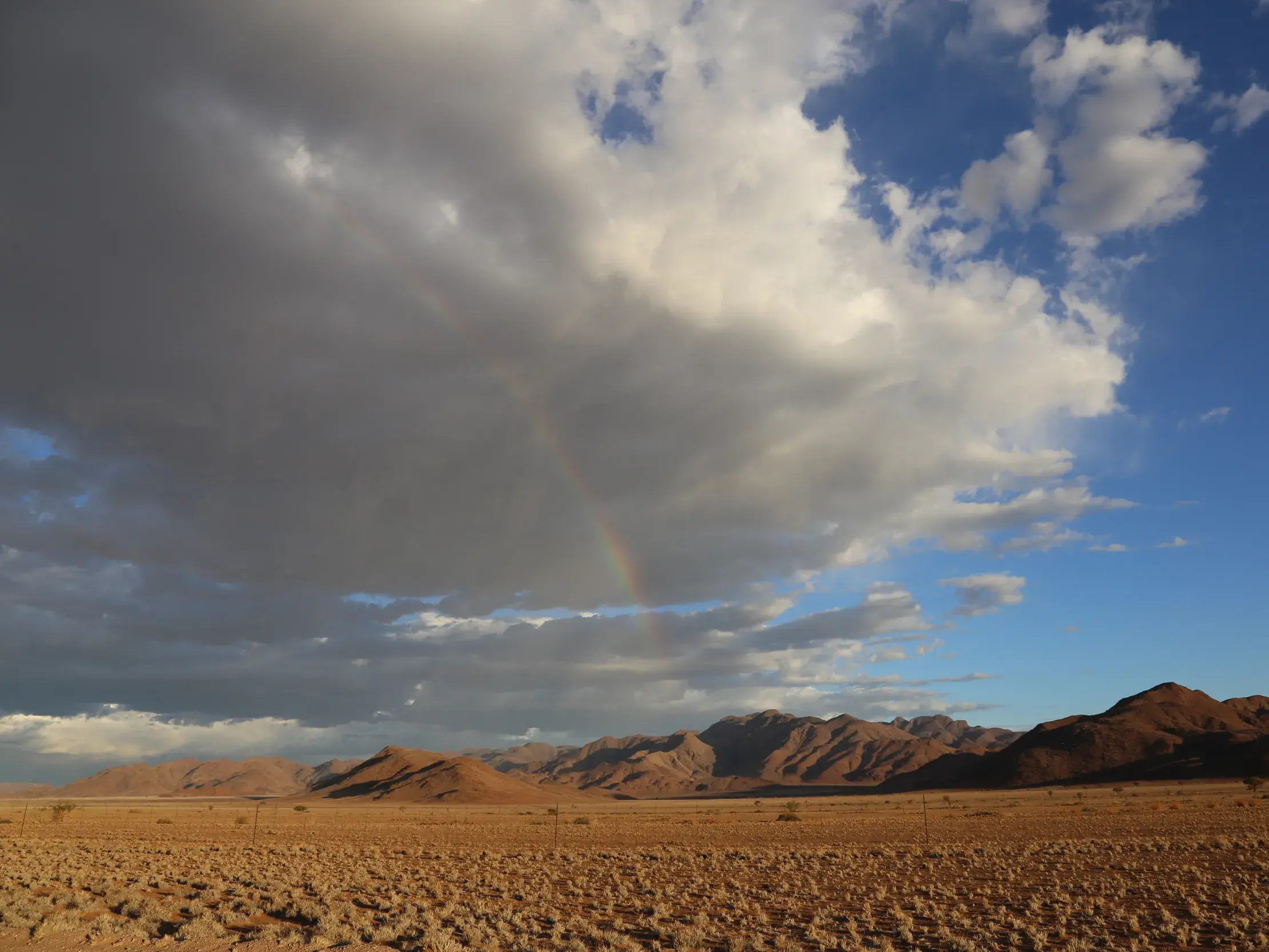 Ein Regenbogen spannt sich unter dramatischen Wolken über die trockene Wüstenlandschaft Namibias mit einer Bergkette am Horizont.