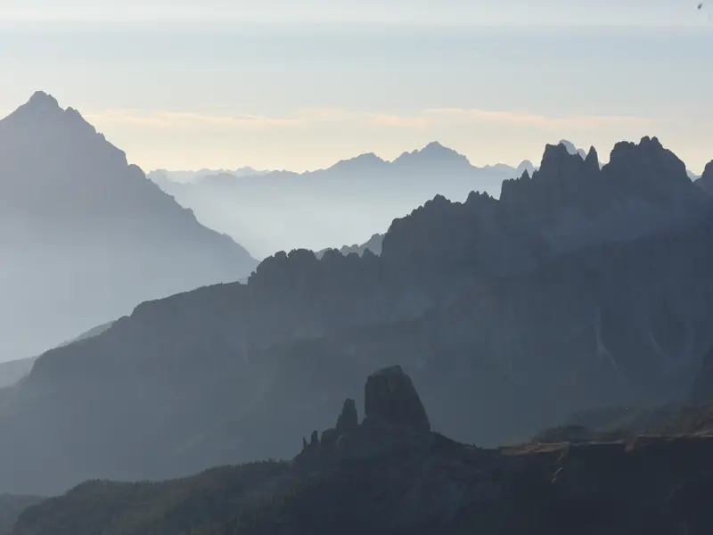 Weitblick beim Trekking auf dem Dolomitenhöhenweg Alta Via 1 über gestaffelte, neblige Berggipfel bei Sonnenaufgang.