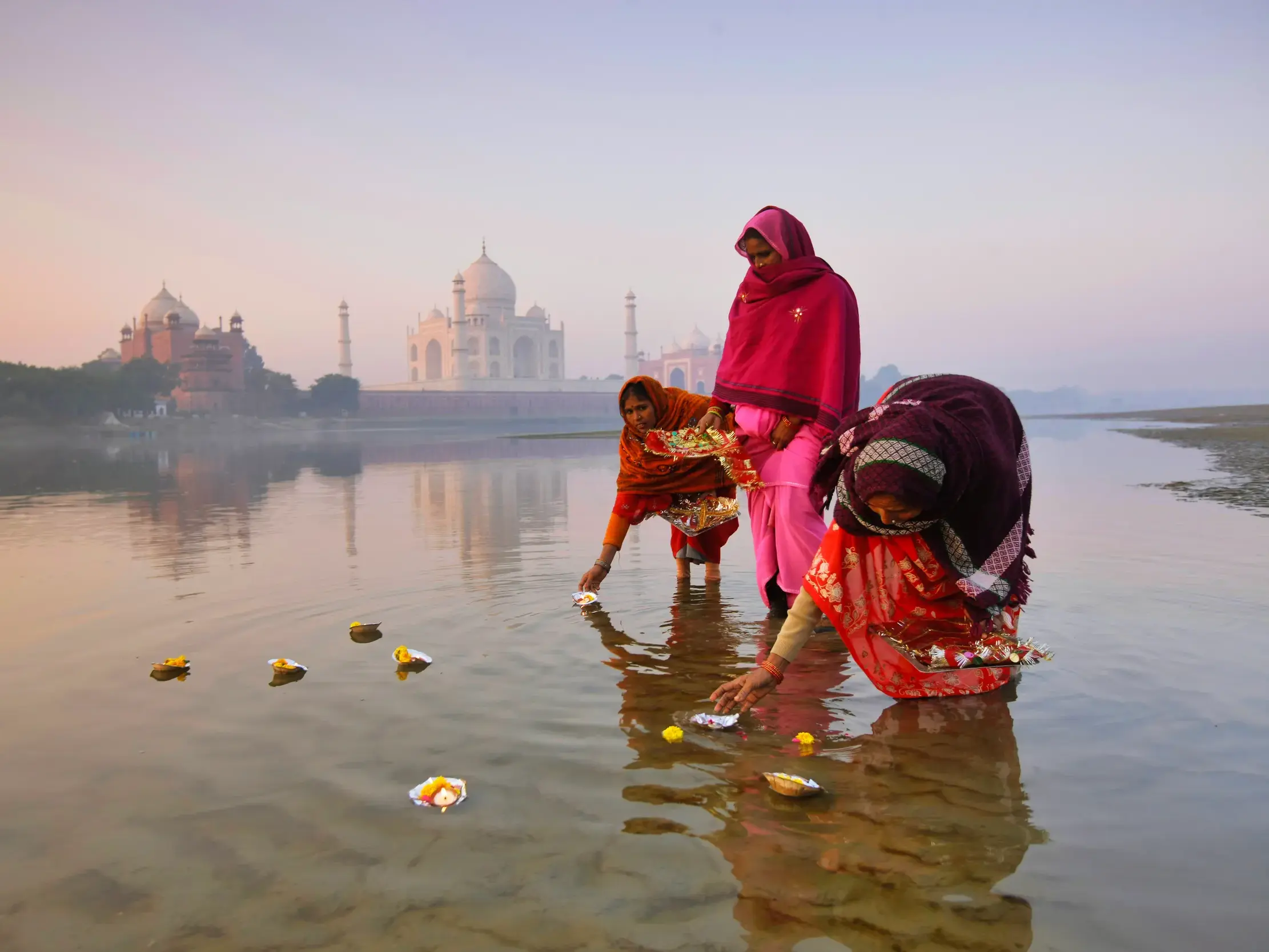 Drei Frauen in bunten Saris legen bei einer Zeremonie im Yamuna-Fluss Opfergaben ins Wasser, das Taj Mahal im Morgennebel.