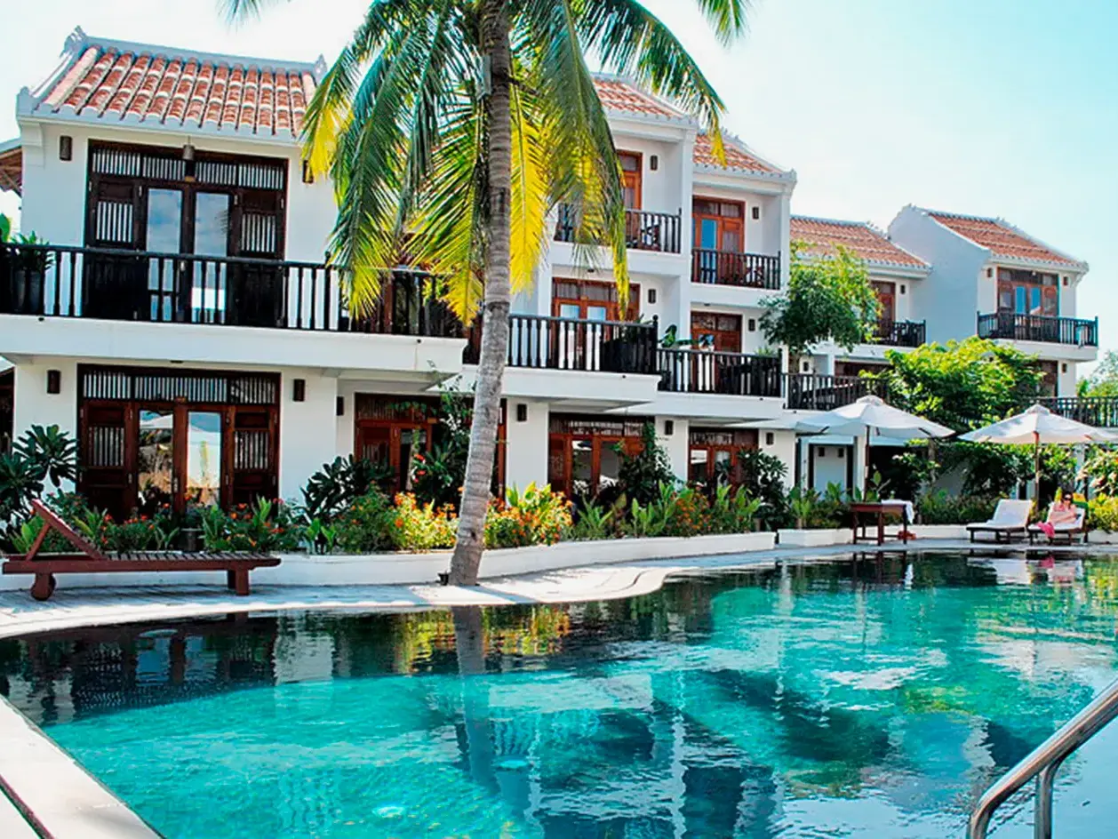 Sonnige Poolanlage des Hotel Coco River Resort in Hoi An mit weißen Gebäuden, Palmen und türkisfarbenem Wasser.