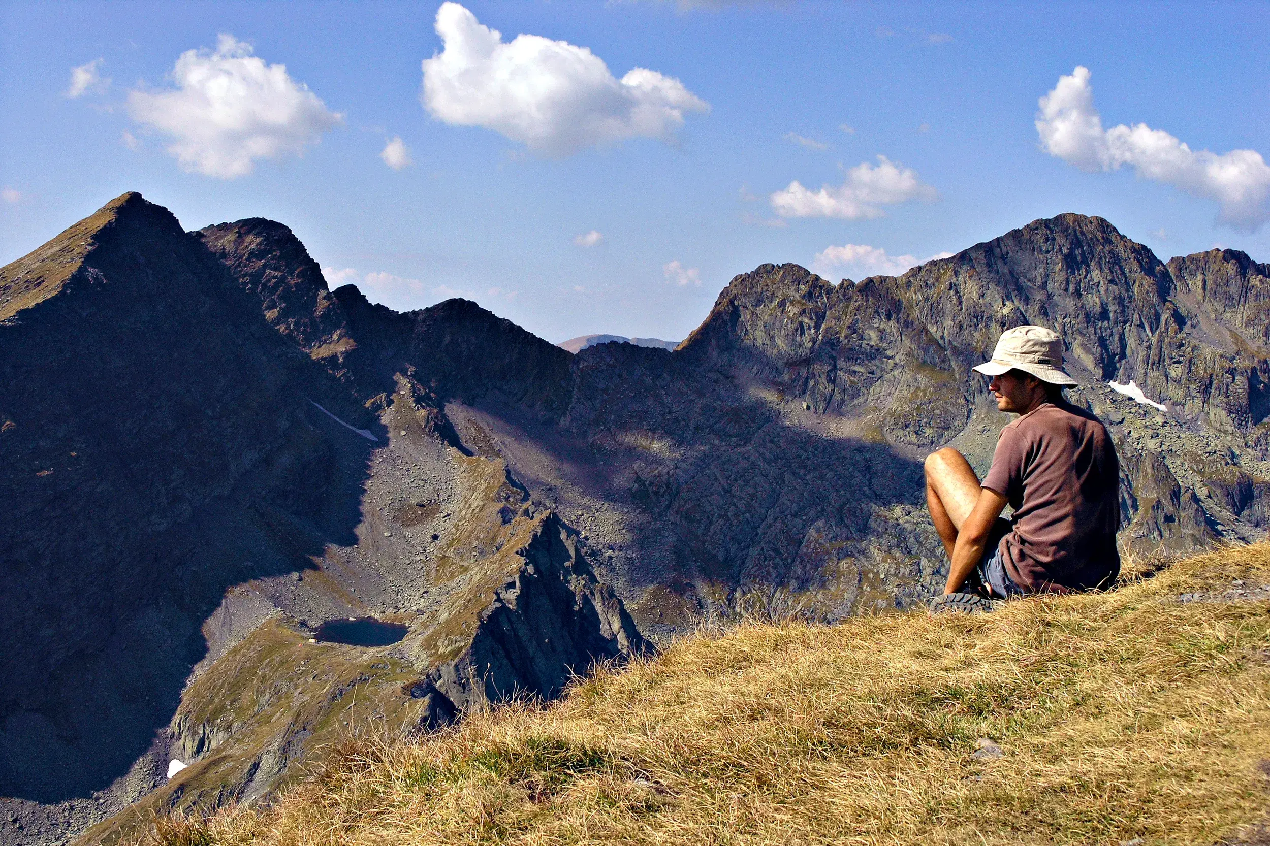 Ein Wanderer mit Hut macht eine Pause auf einem Grashang und blickt über die schroffen Gipfel des Fagaras Gebirges in Rumänien.