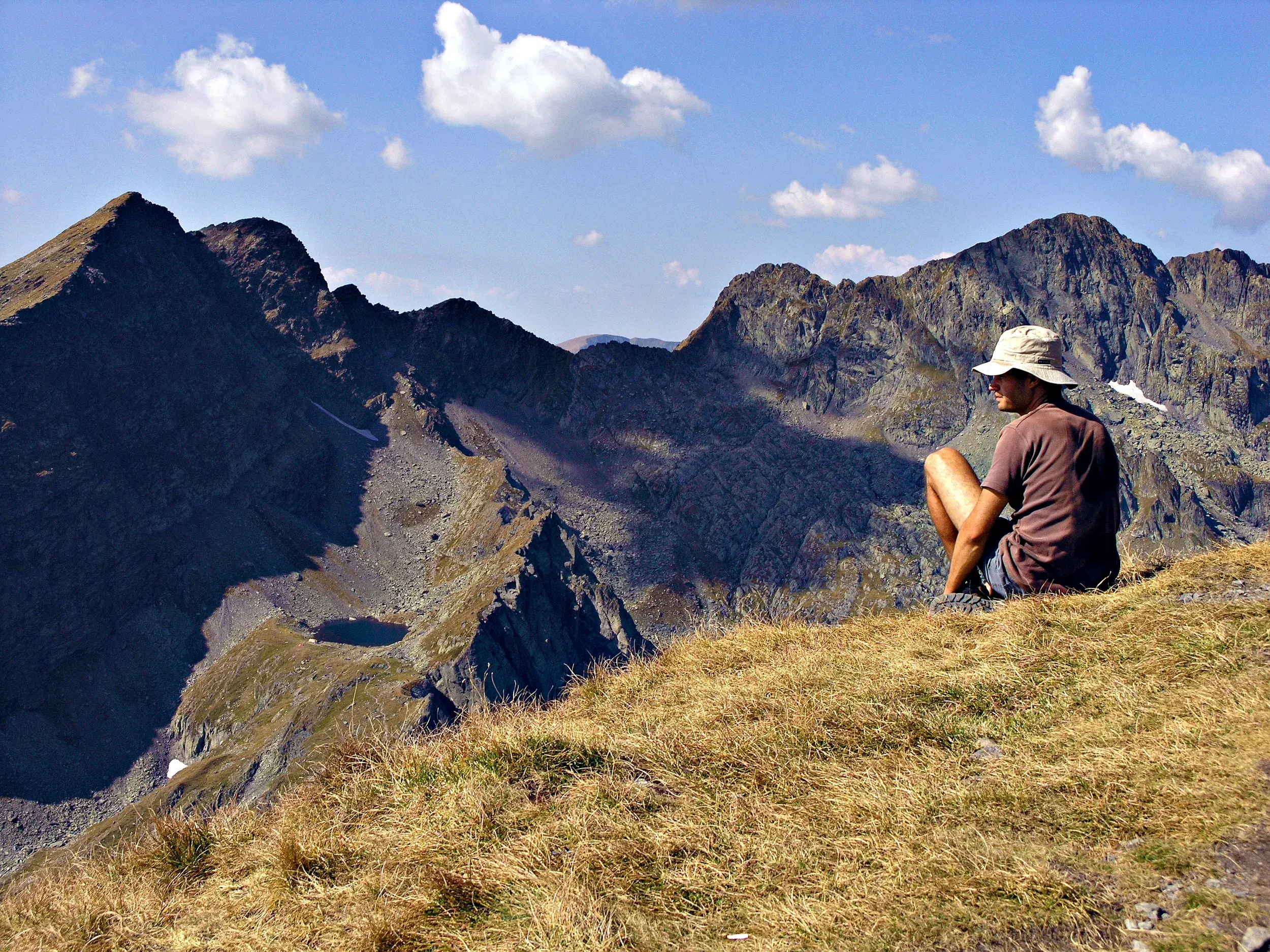 Ein Wanderer mit Hut macht eine Pause auf einem Grashang und blickt über die schroffen Gipfel des Fagaras Gebirges in Rumänien.