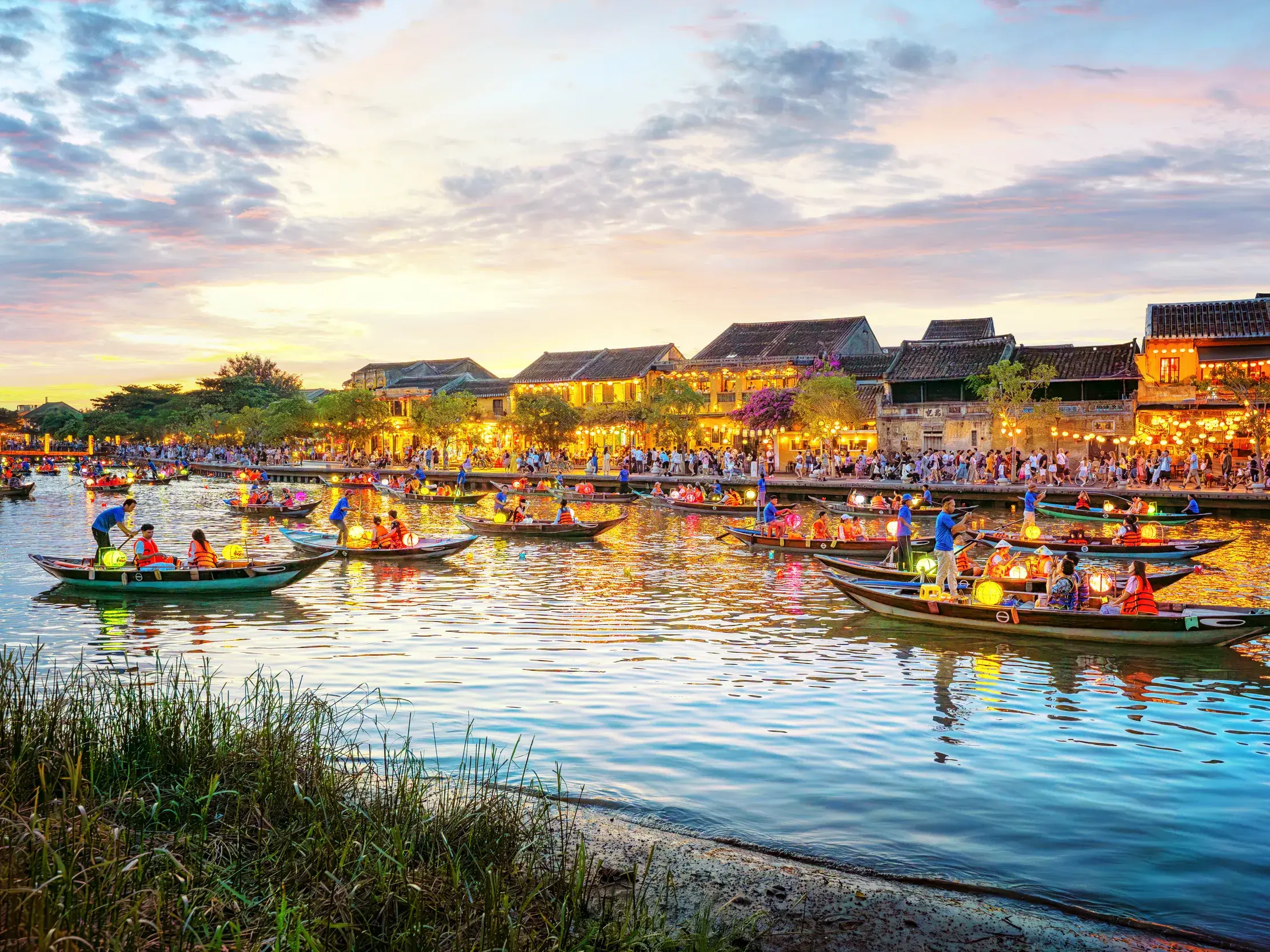 Zahlreiche Boote mit bunten Lampions gleiten bei Dämmerung über den Fluss in der Altstadt von Hoi An, einem UNESCO-Welterbe.