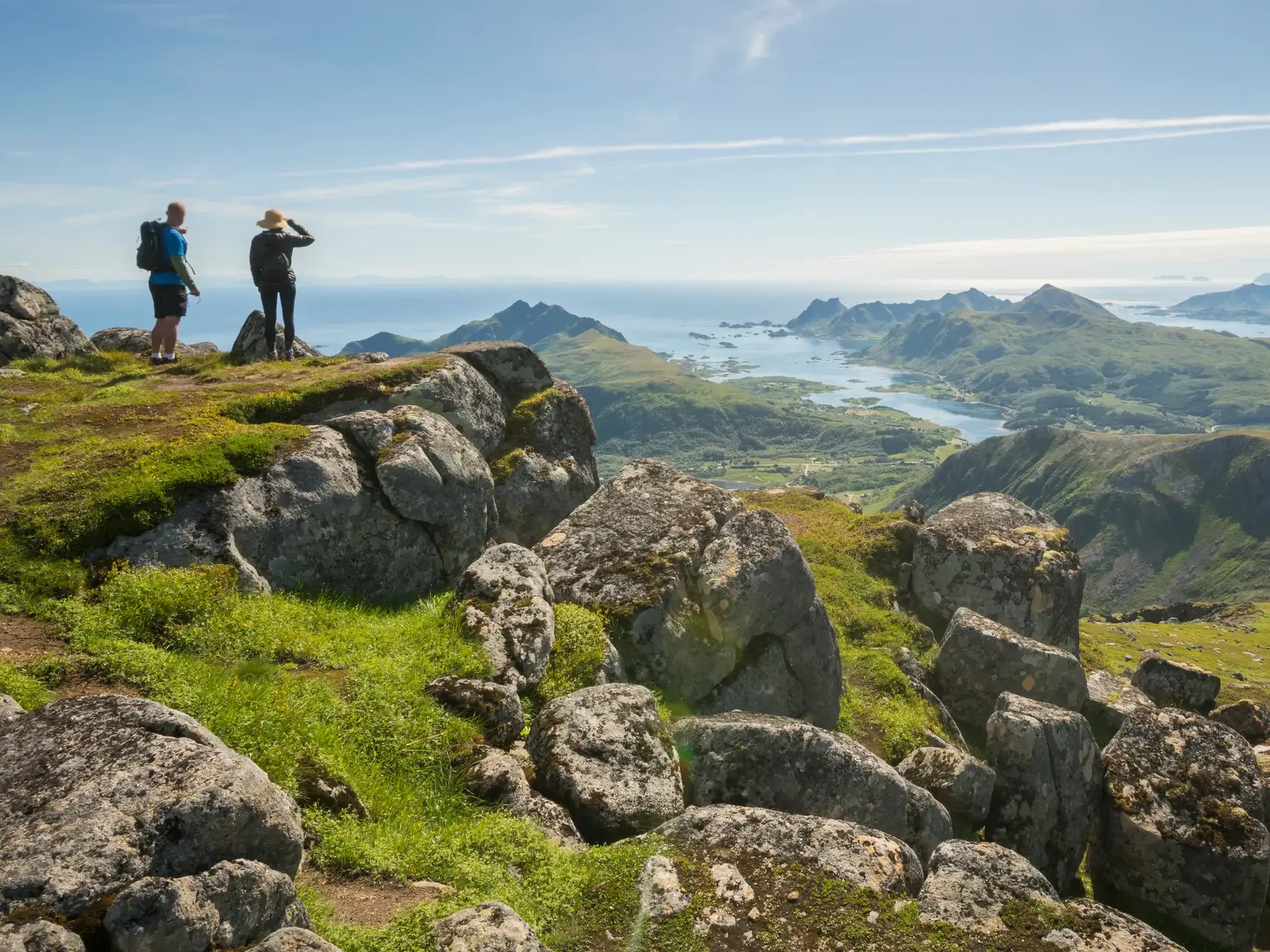 Zwei Wanderer genießen bei einer Trekking-Tour auf einem Gipfel der Lofoten den weiten Ausblick über die Fjordlandschaft Norwegens.