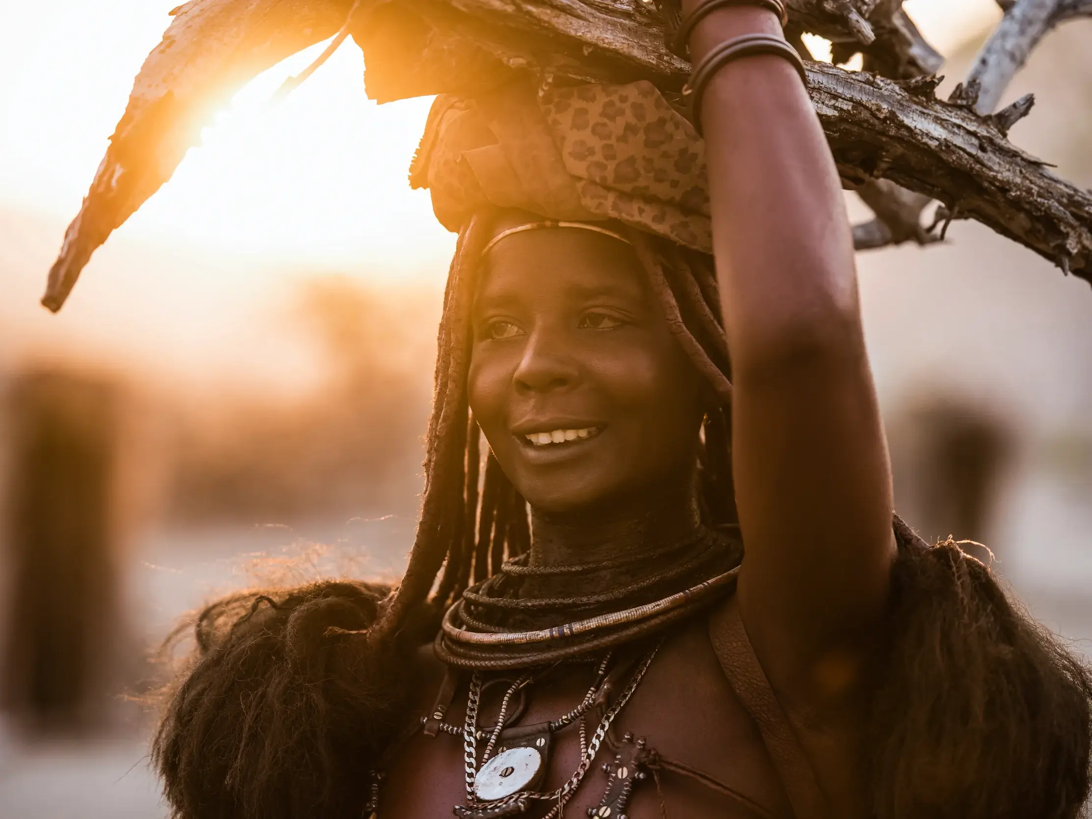 Lächelnde Himba-Frau in Namibia mit traditionellem Schmuck balanciert ein Stück Holz auf dem Kopf im warmen Licht des Sonnenuntergangs.