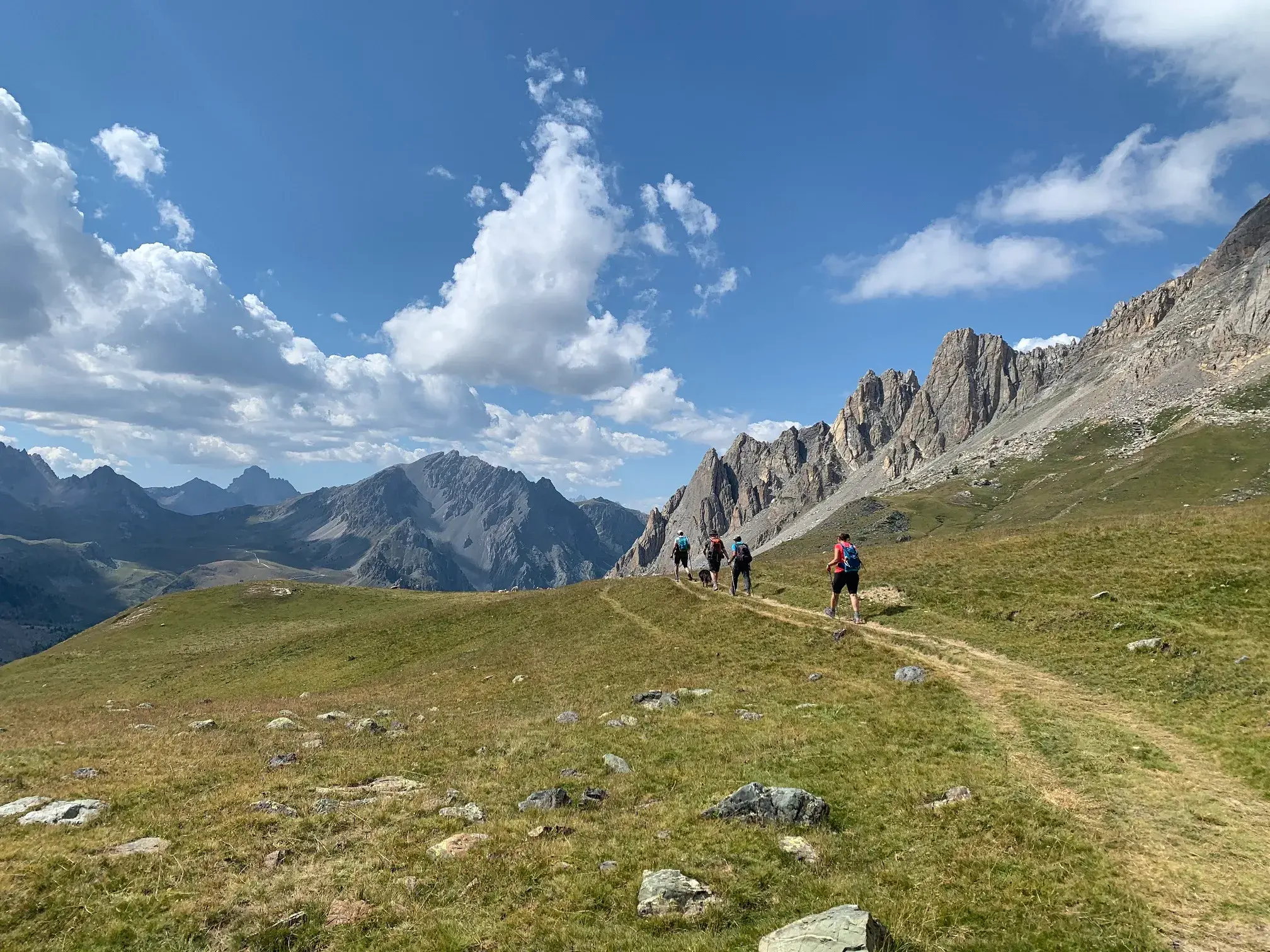 Eine kleine Wandergruppe auf einem schmalen Pfad durch Almwiesen mit Panoramablick auf die Alpengipfel im Maira-Tal.