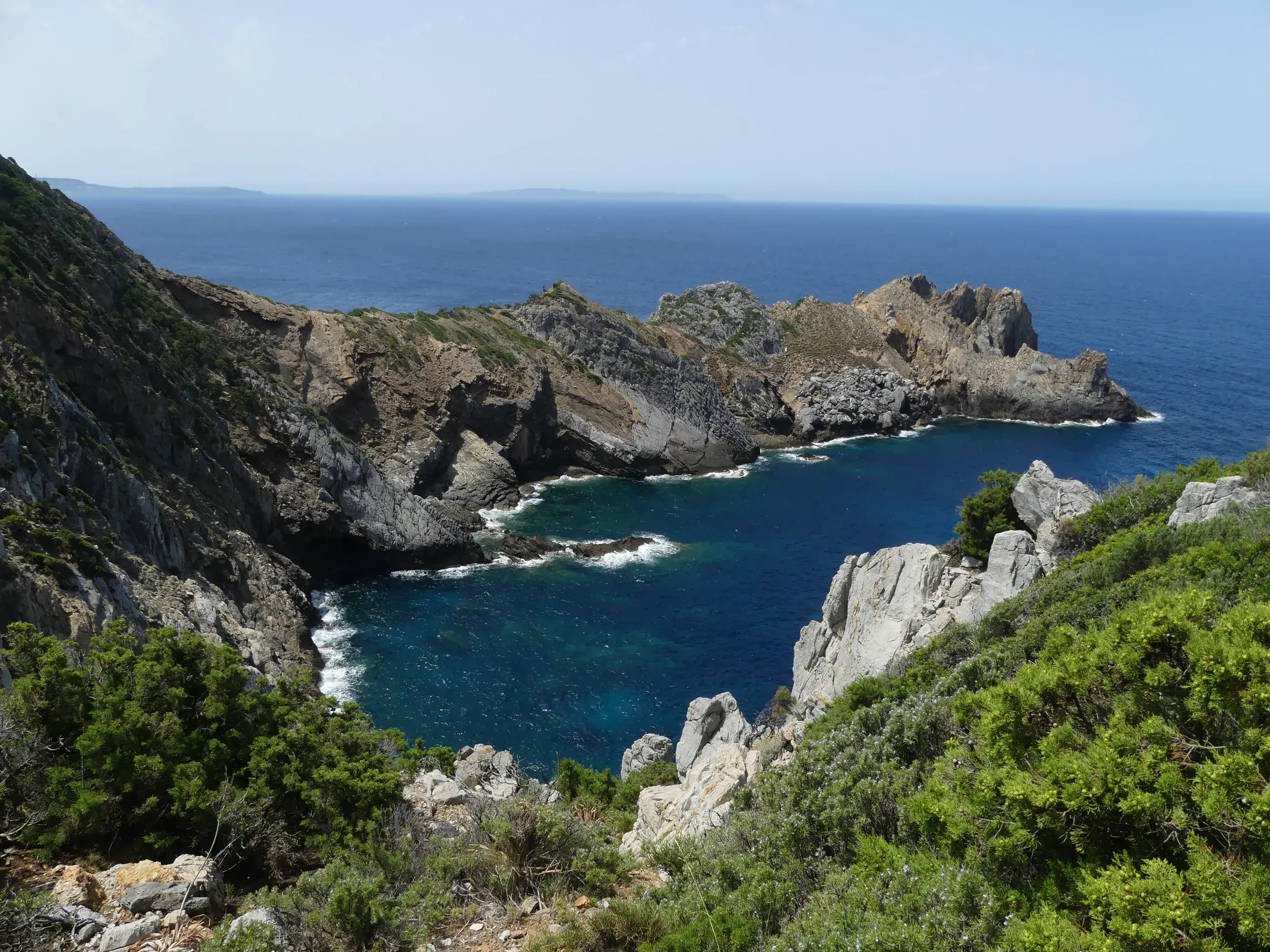 Blick von einem Wanderweg auf die zerklüftete Felsküste und eine tiefblaue Bucht am Capo Pecora, Sardinien.