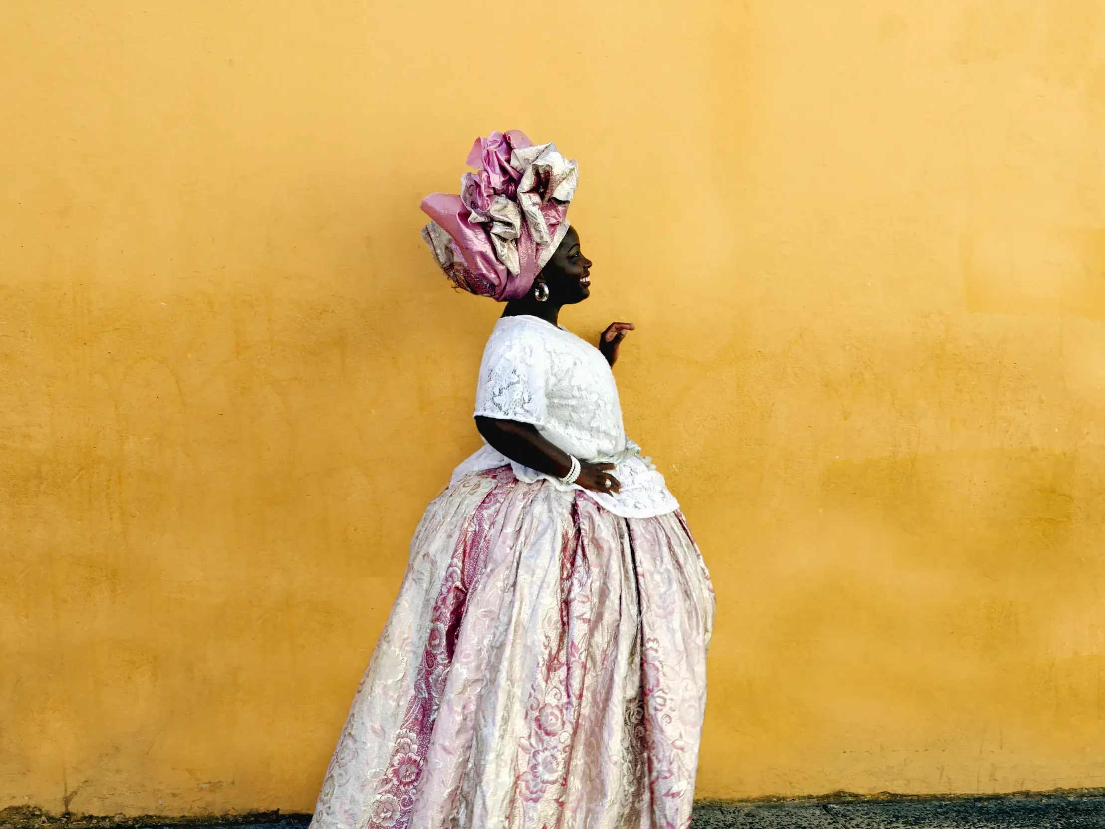 Eine Baiana in prachtvoller, weiß-rosa Tracht und Turban steht lächelnd im Profil vor einer sonnengelben Wand in Salvador.