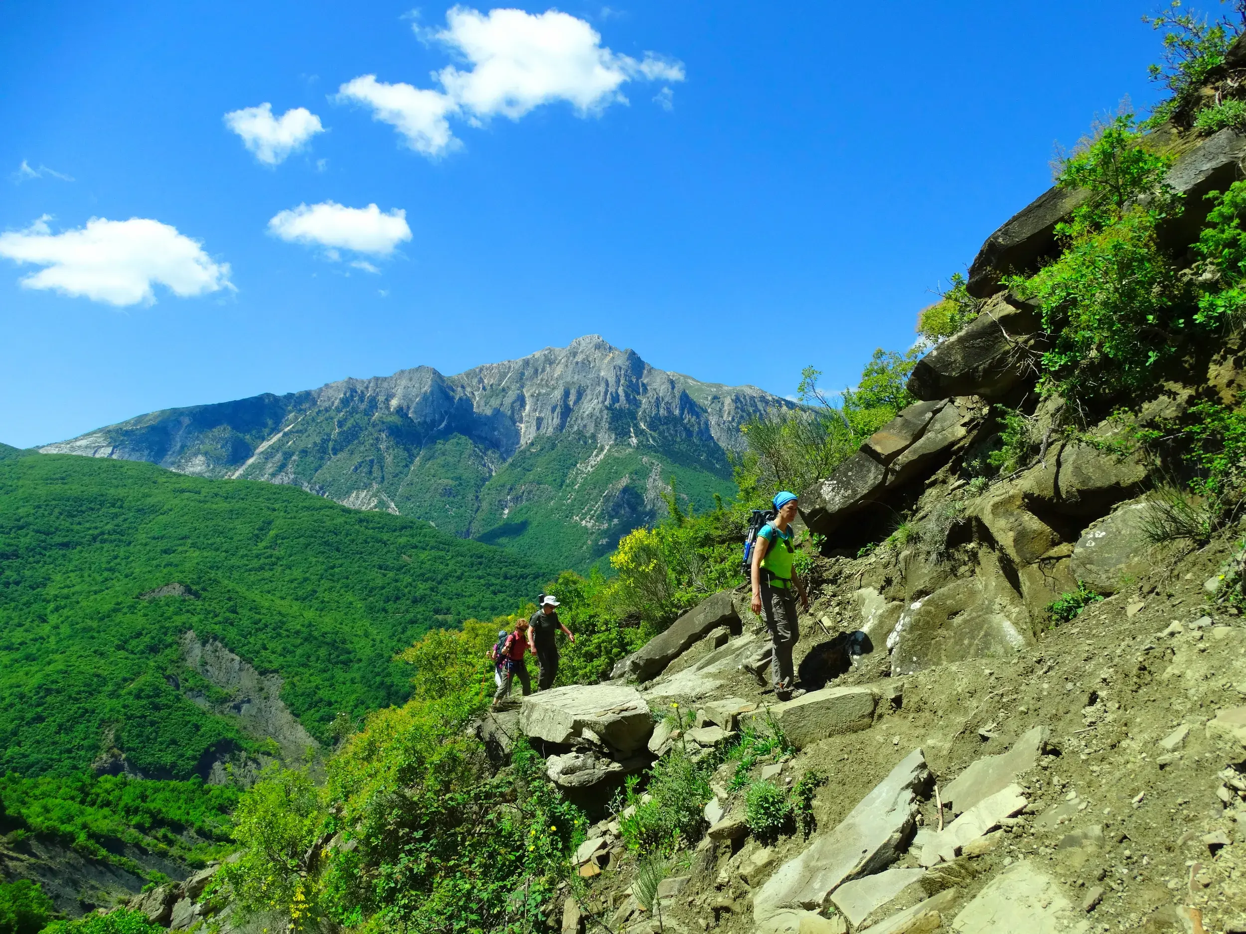 Eine Wandergruppe beim Trekking auf einem steinigen Pfad im grünen Küstengebirge Albaniens mit Blick auf einen Gipfel.