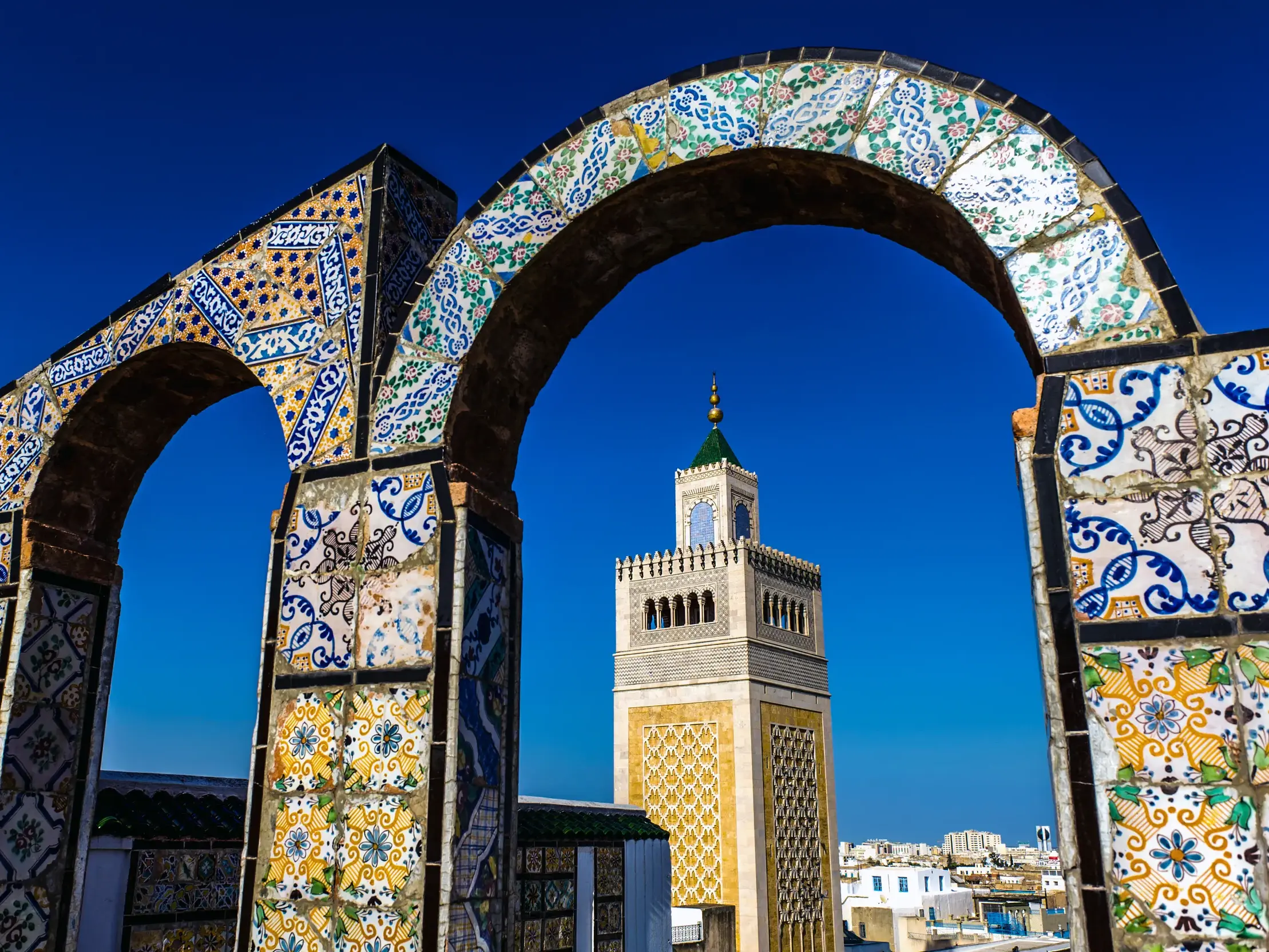 Ein mit Mosaikfliesen verzierter Torbogen rahmt das Minarett der Ez-Zitouna-Moschee vor strahlend blauem Himmel in Tunis ein.