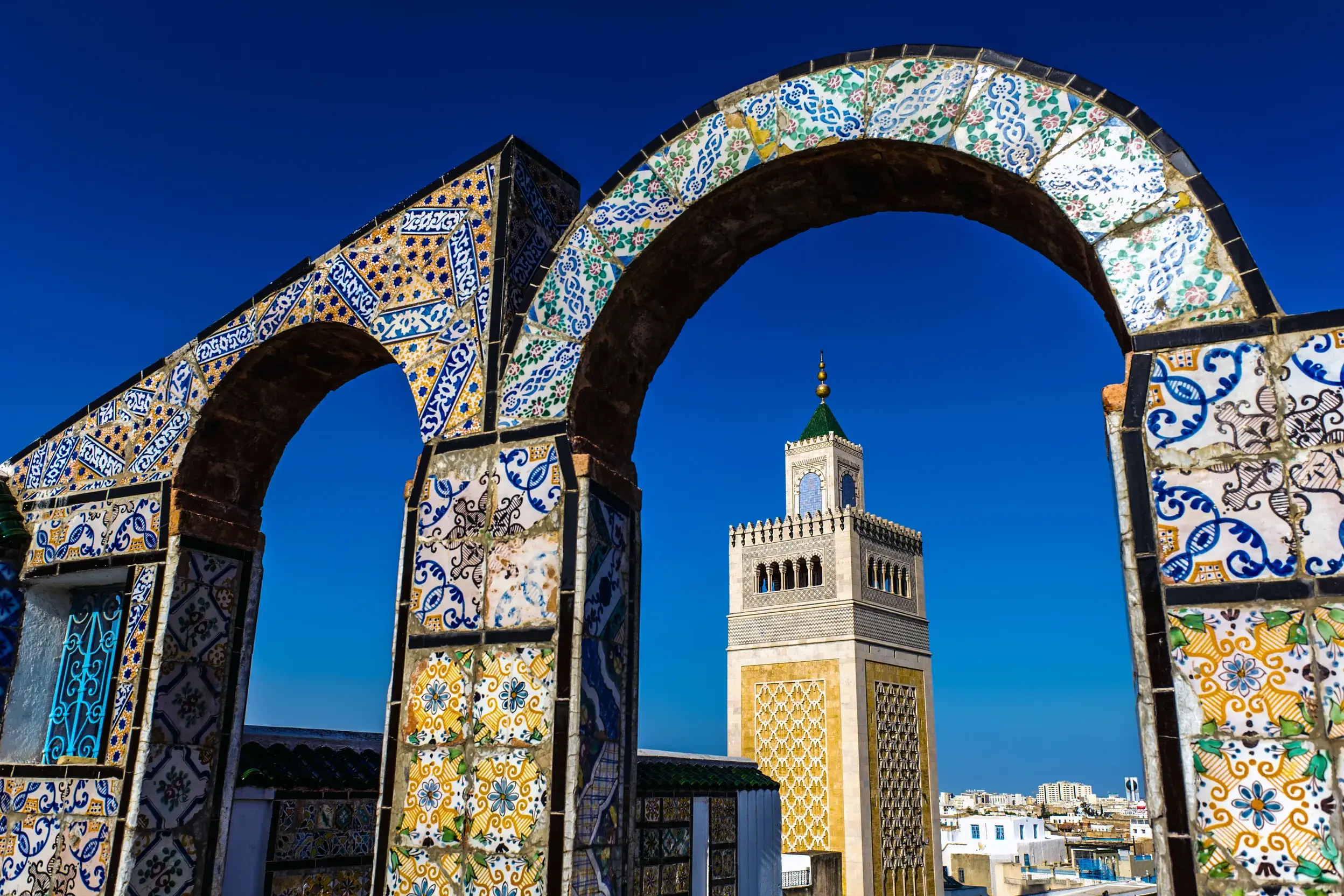 Ein mit Mosaikfliesen verzierter Torbogen rahmt das Minarett der Ez-Zitouna-Moschee vor strahlend blauem Himmel in Tunis ein.