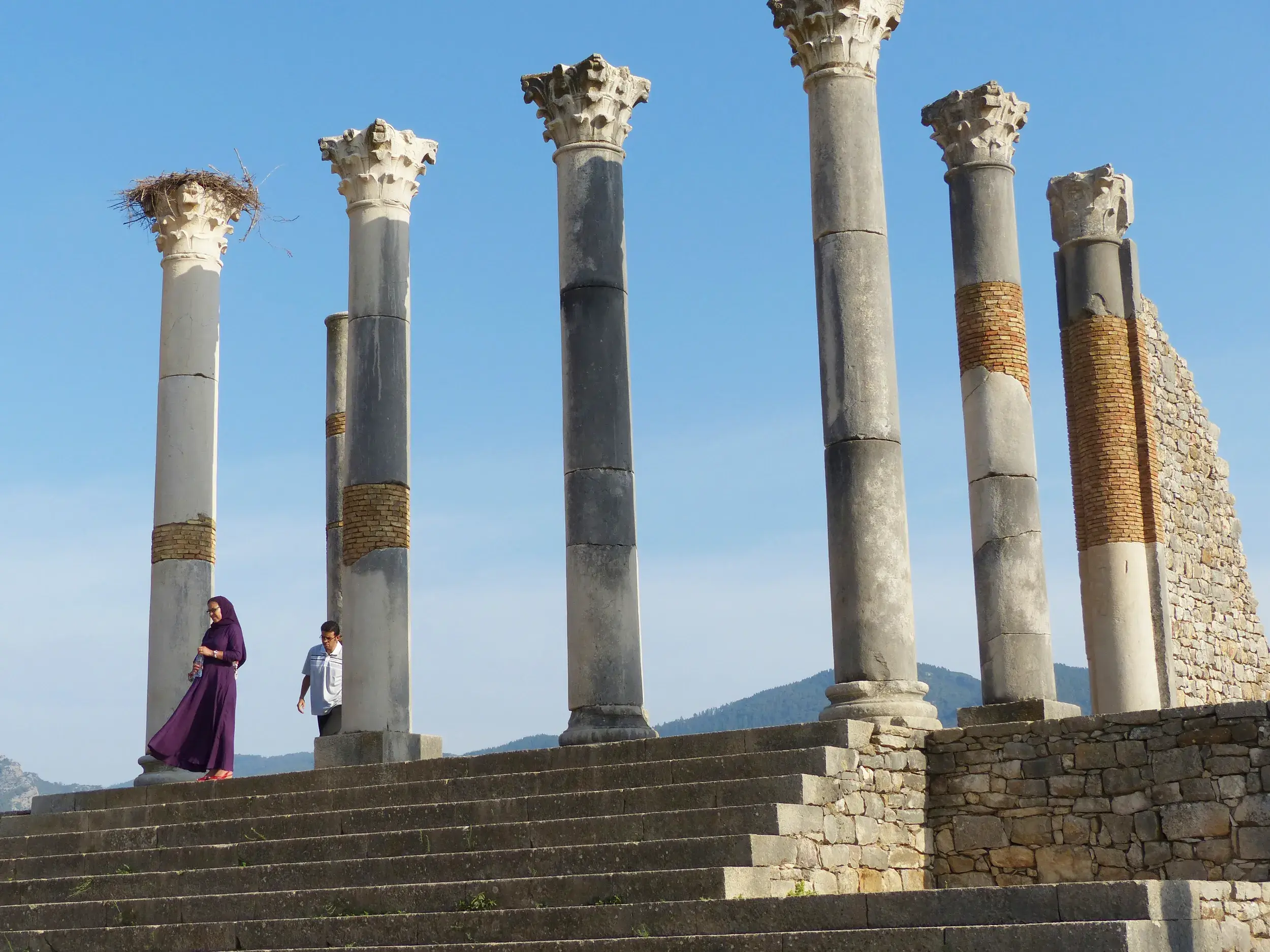 Hohe römische Säulen mit Storchennest in der Ruinenstadt Volubilis, davor eine Frau in lila Gewand auf Steintreppen.