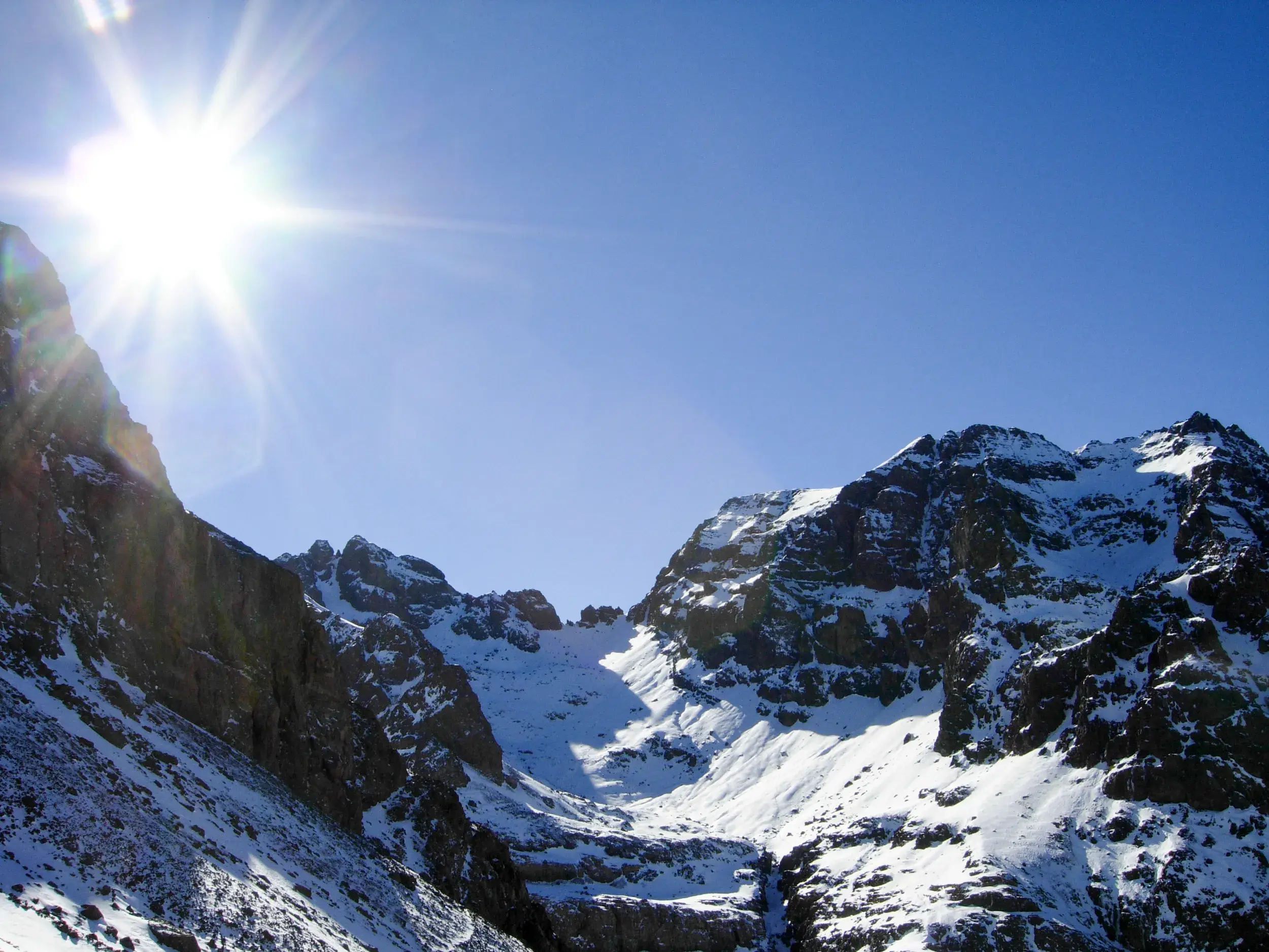 Strahlender Sonnenschein über der schroffen, schneebedeckten Berglandschaft des Hohen Atlas in Marokko bei klarem Himmel.