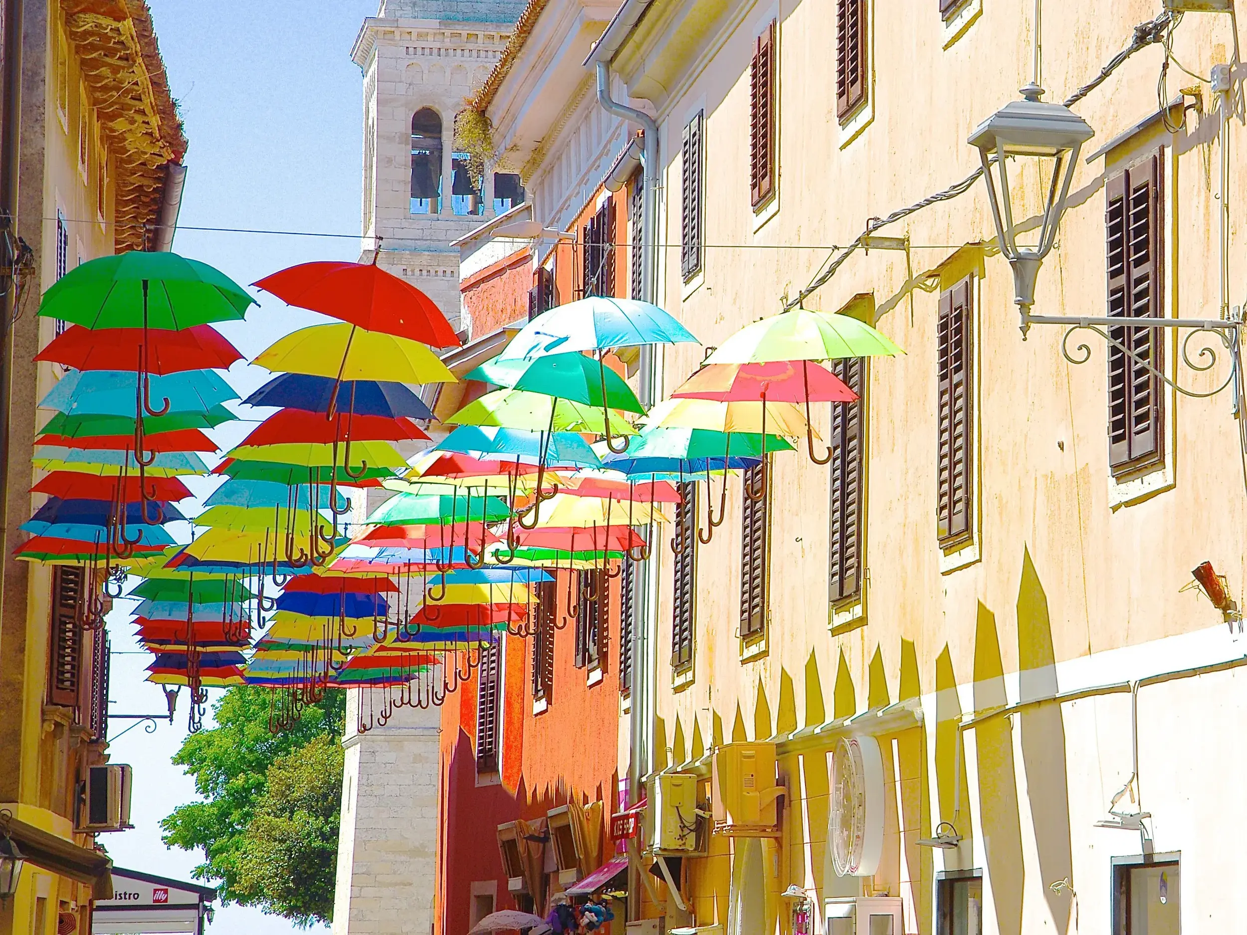 Farbenfrohe Regenschirme schweben als Kunstinstallation über einer sonnigen Gasse in der Altstadt von Novigrad in Istrien.