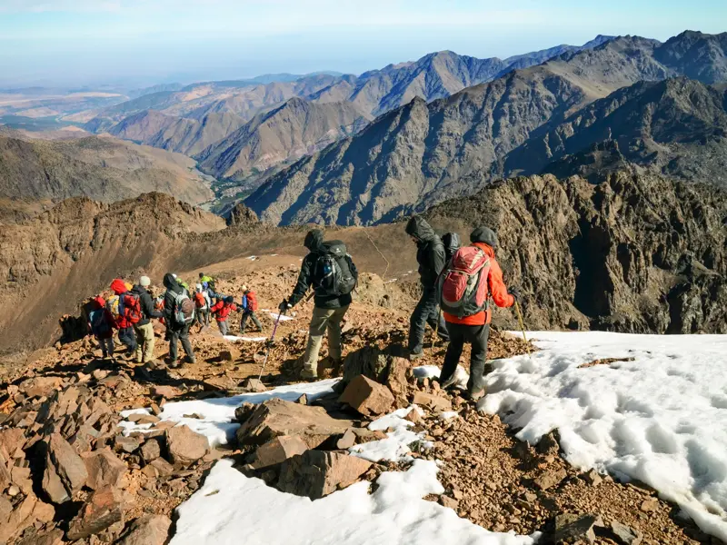 Eine Trekkinggruppe wandert auf einem felsigen, teilweise schneebedeckten Grat während der Besteigung des Djebel Toubkal im Hohen Atlas.