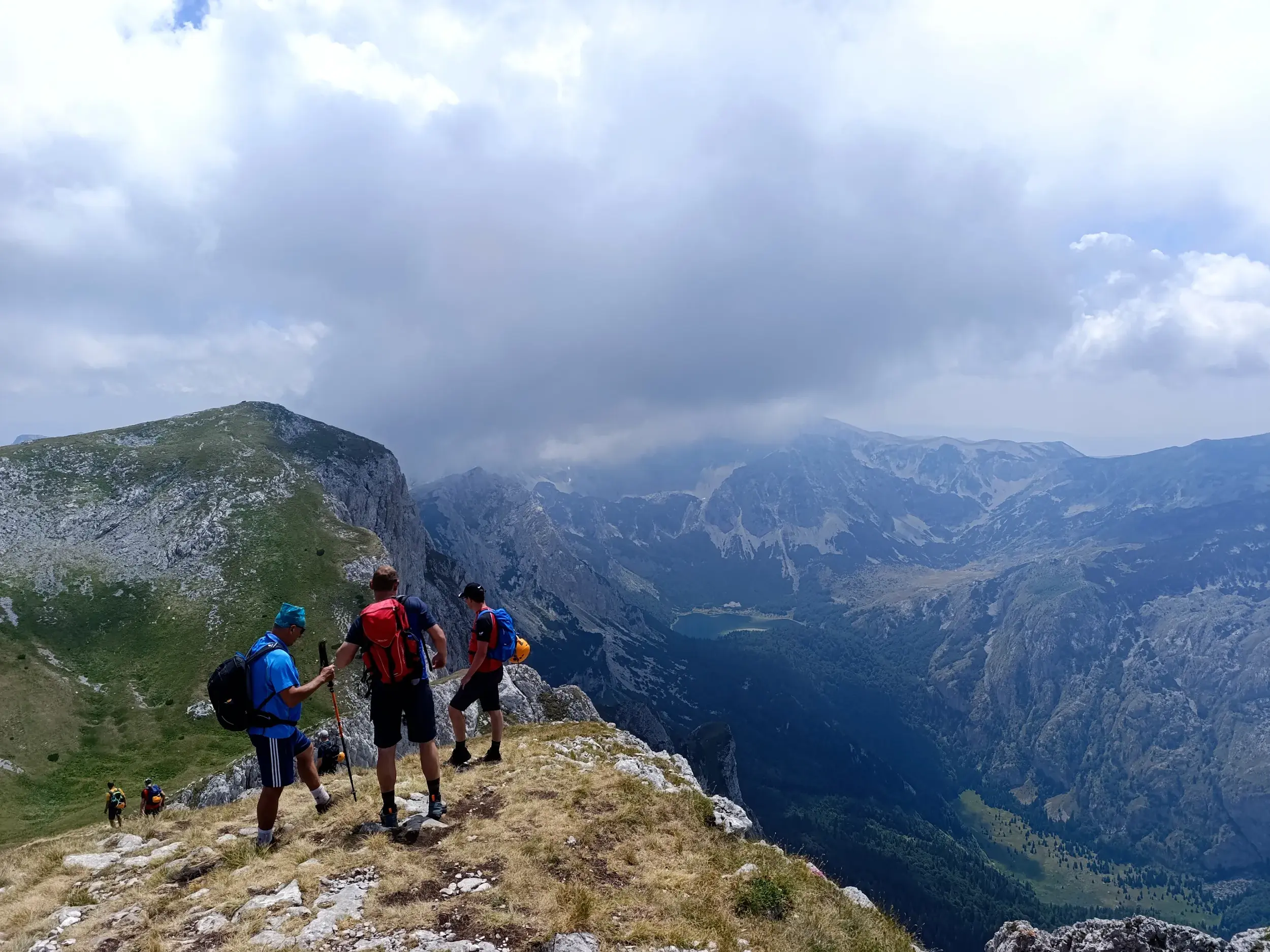 Drei Wanderer mit Rucksäcken auf einem grasbewachsenen Grat blicken über ein tiefes Tal und eine weite Gebirgslandschaft.