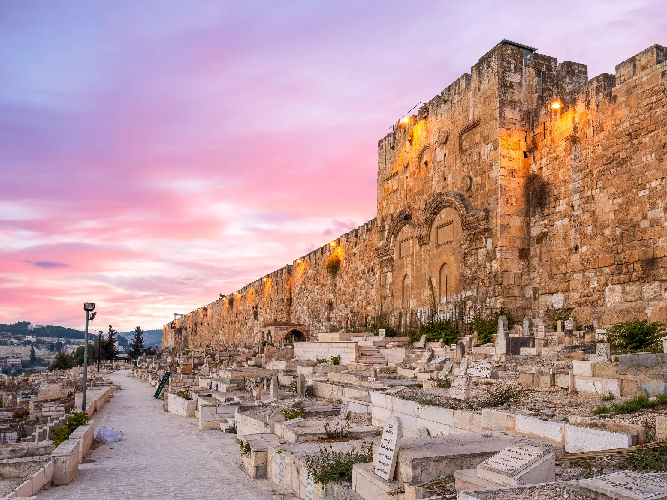 Die historische Stadtmauer von Jerusalem mit dem Goldenen Tor, beleuchtet im Abendrot, davor ein jüdischer Friedhof.