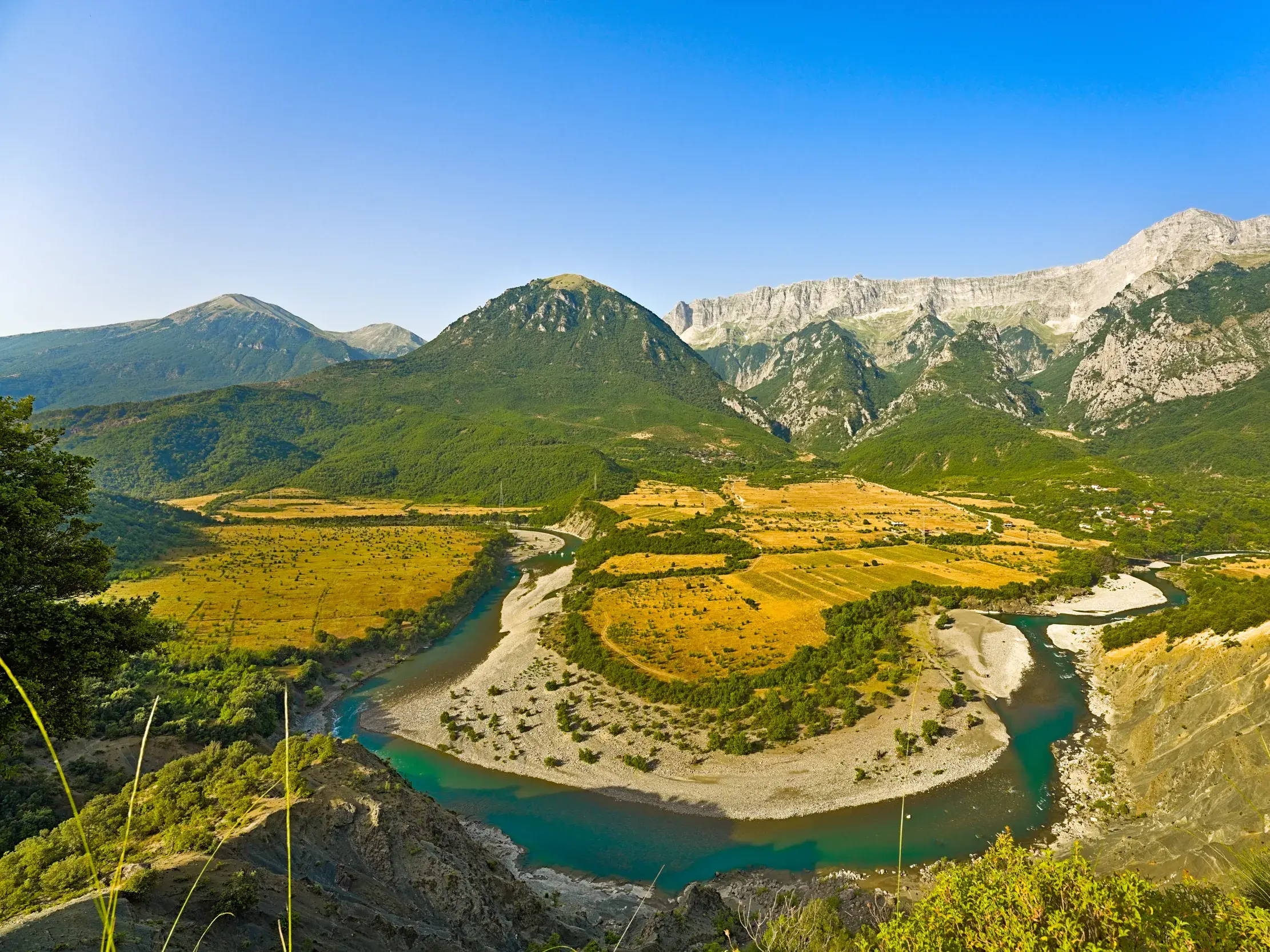 Die naturbelassene, türkisblaue Vjosa mäandert durch ein weites Tal in der albanischen Gebirgslandschaft bei strahlend blauem Himmel.