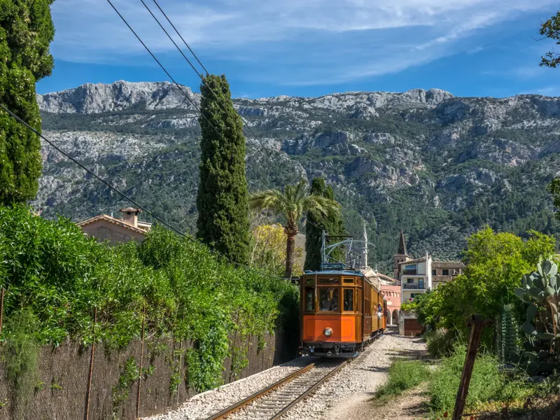 Historische, orange Trambahn von Sóller auf dem Weg durch eine mediterrane Landschaft vor dem Tramuntana-Gebirge auf Mallorca.