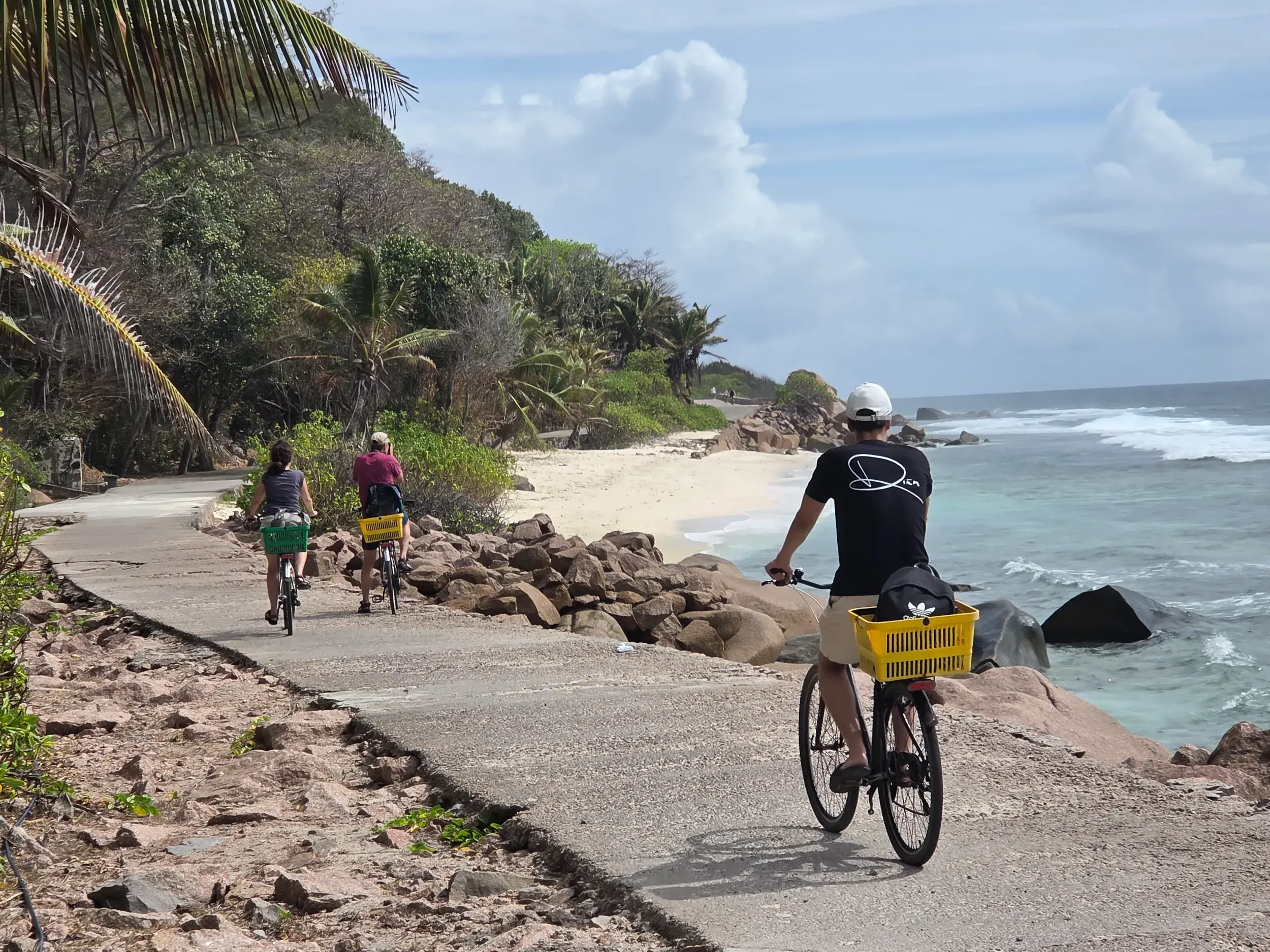 Eine kleine Reisegruppe radelt auf einem Küstenweg auf La Digue, vorbei an Granitfelsen und einem weißen Sandstrand.