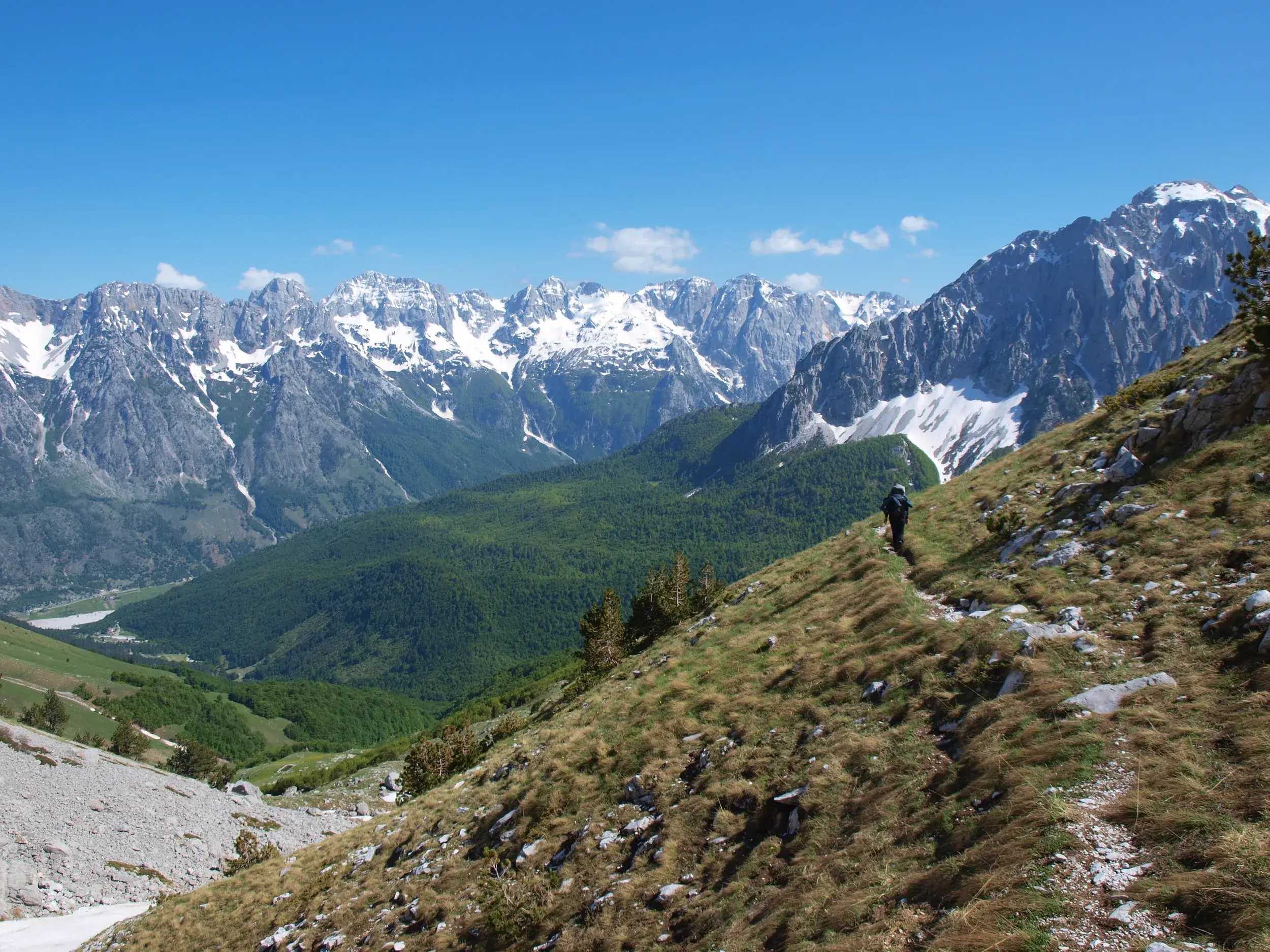 Ein Wanderer folgt einem schmalen Pfad über einen Grashang mit Panoramablick auf das Valbona-Tal und die albanischen Alpen.
