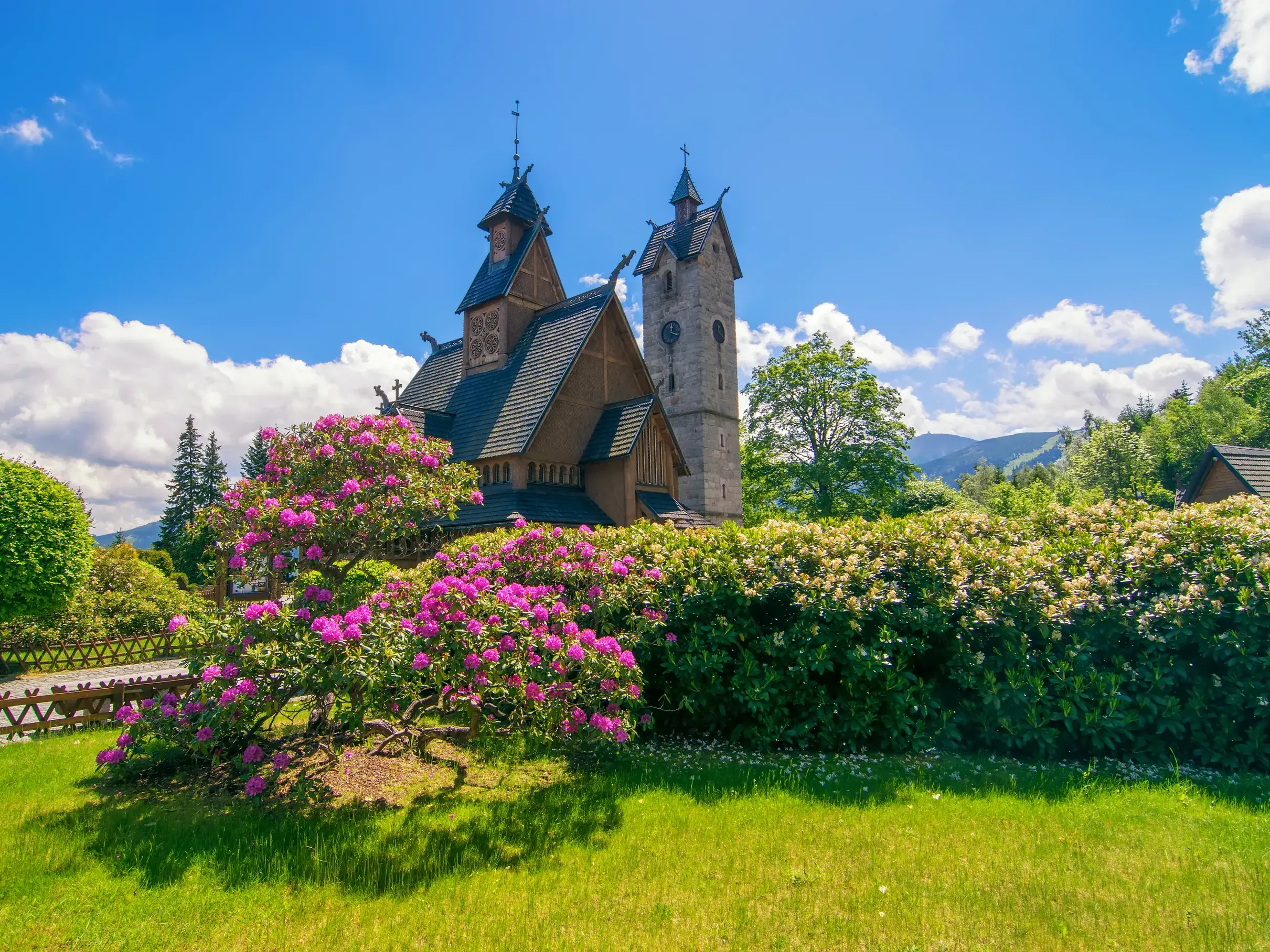 Die mittelalterliche Stabkirche Wang bei Karpacz in Polen mit blühendem Rhododendron vor der Bergkulisse des Riesengebirges.