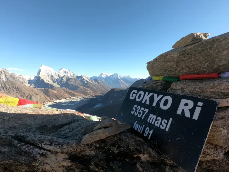 Schild am Gipfel des Gokyo Ri mit Panoramablick auf die schneebedeckten Himalaya-Gipfel und den türkisen Gokyo-See.
