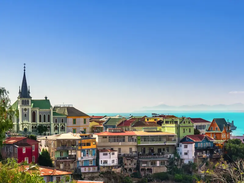 Panoramablick auf die bunten Häuser und eine Kirche der UNESCO-Hafenstadt Valparaíso in Chile am leuchtend blauen Meer.