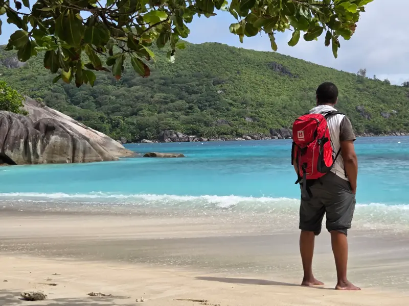 Wanderer mit rotem Hauser-Rucksack am Sandstrand auf den Seychellen blickt auf Granitfelsen und das türkise Meer.