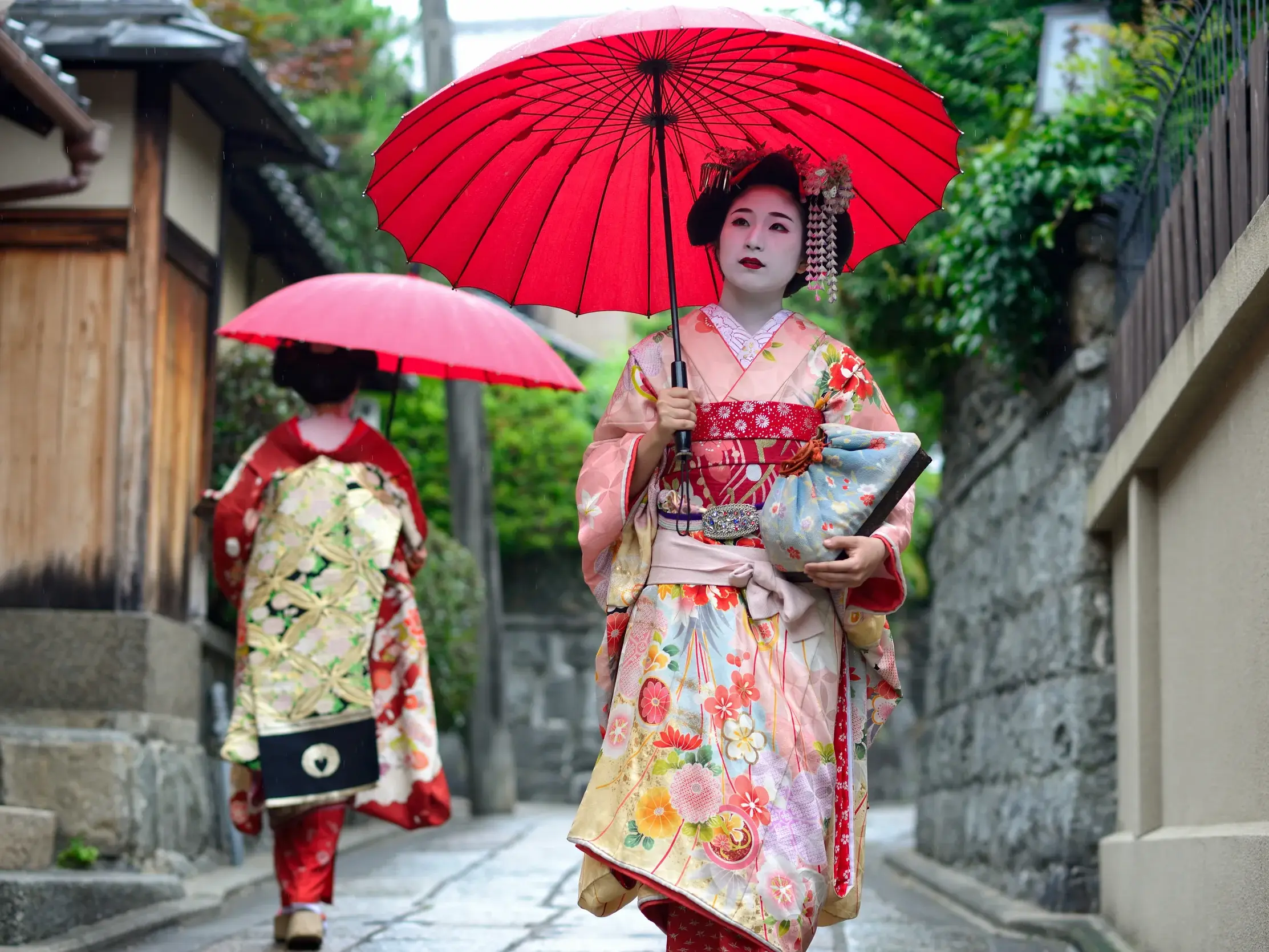 Zwei Geishas in farbenfrohen Kimonos spazieren mit roten Regenschirmen durch eine historische Gasse in Kyoto.