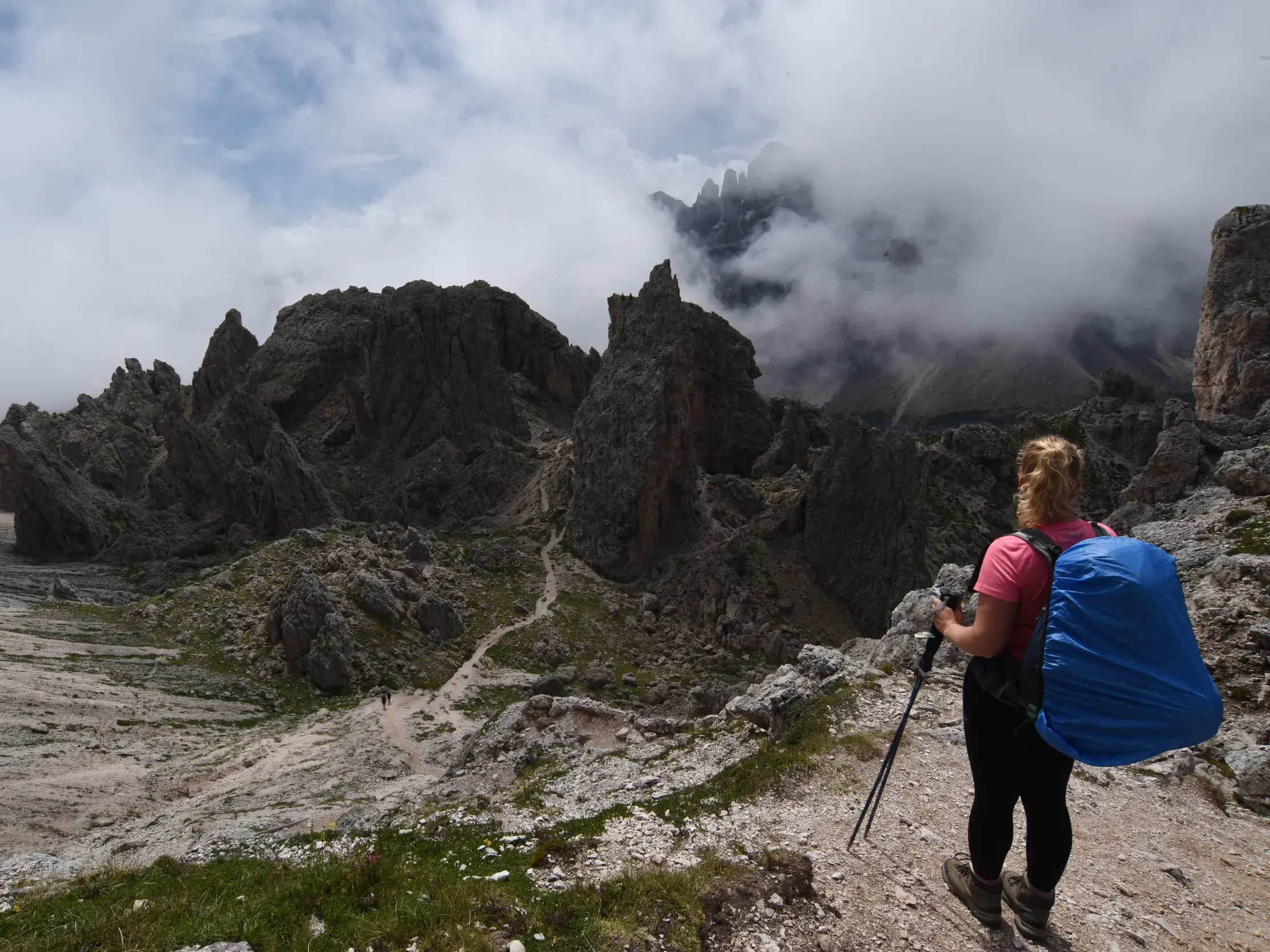 Eine Wanderin mit Rucksack und Stöcken blickt beim Trekking auf die schroffe, wolkenverhangene Sella-Gruppe in den Dolomiten.