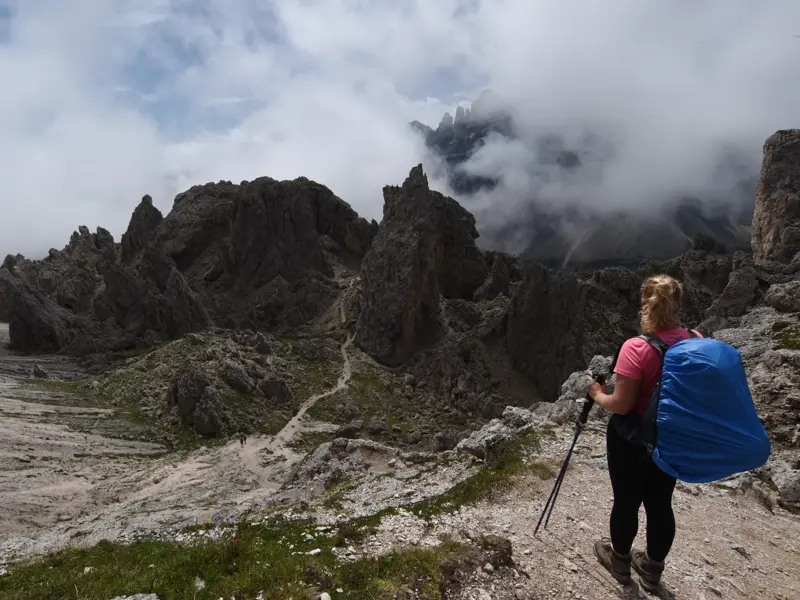 Eine Wanderin mit Rucksack und Stöcken blickt beim Trekking auf die schroffe, wolkenverhangene Sella-Gruppe in den Dolomiten.