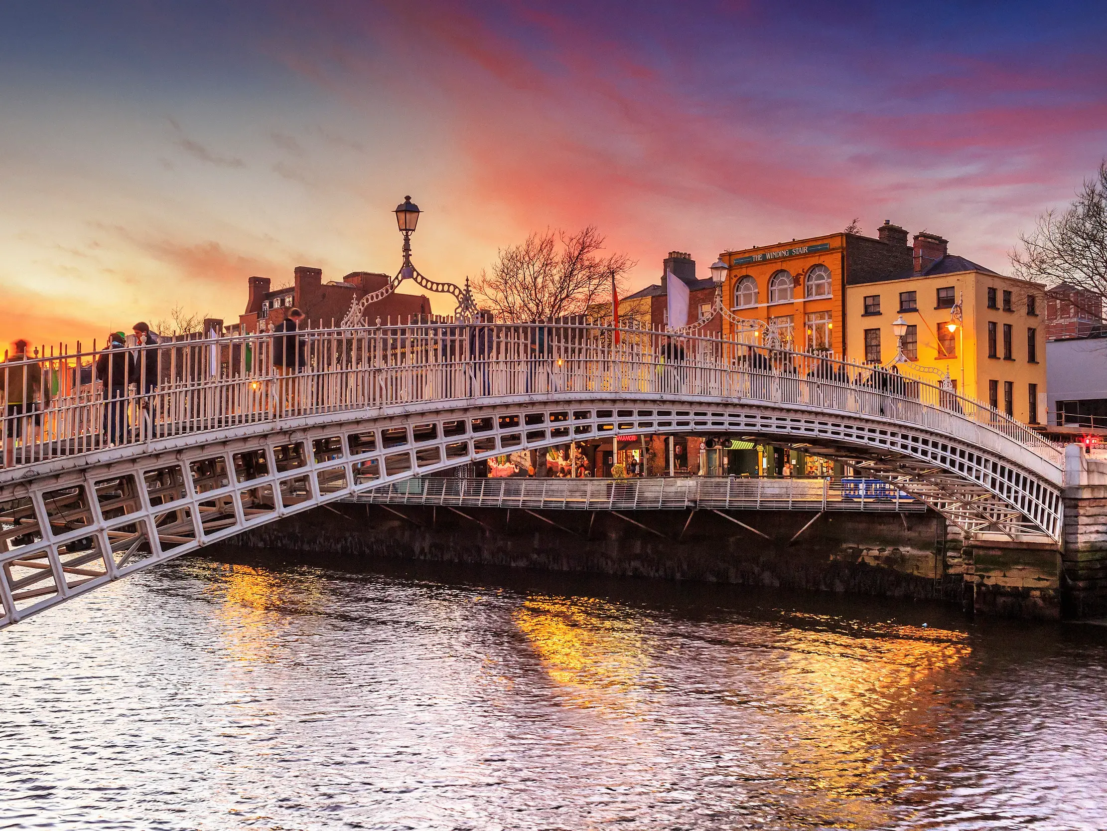 Die weiße, gusseiserne Ha'penny Bridge in Dublin, über die Fußgänger bei malerischem Sonnenuntergang den Fluss Liffey überqueren.