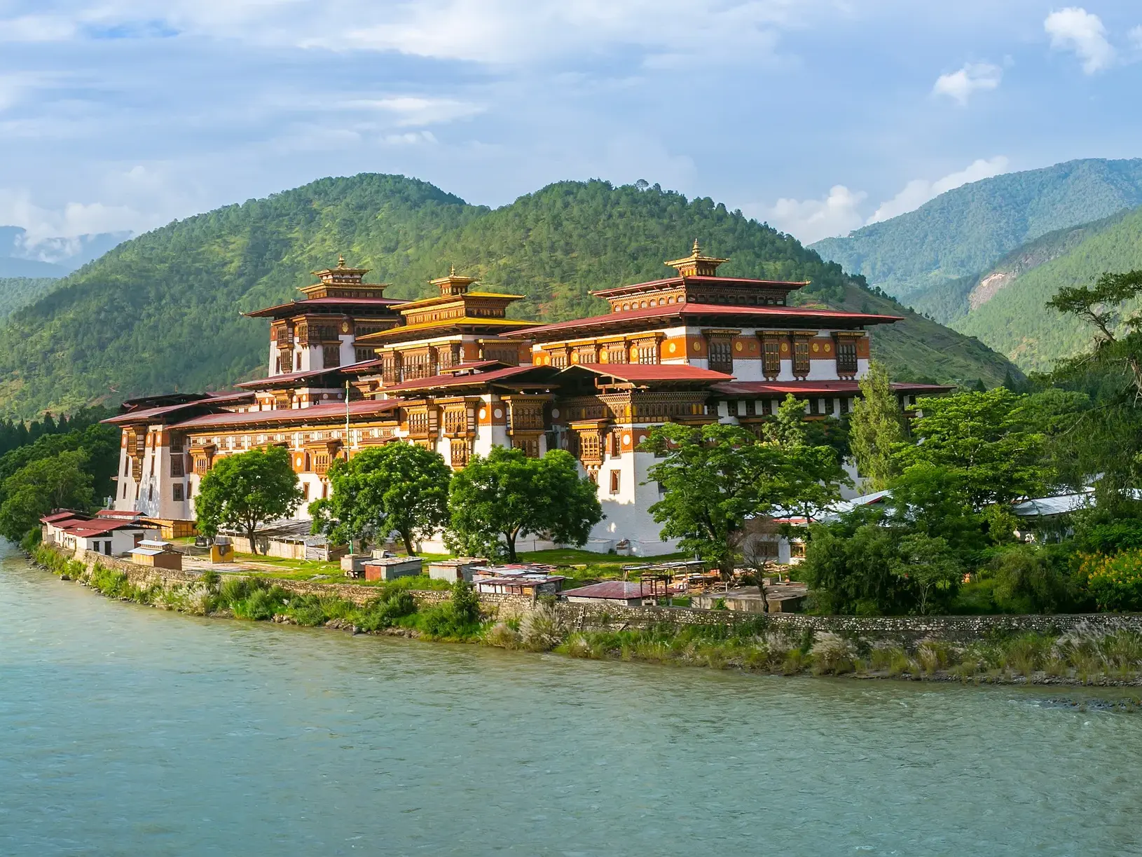 Die eindrucksvolle Klosterburg Punakha Dzong in Bhutan liegt malerisch am Ufer eines Flusses vor grünen Bergen.