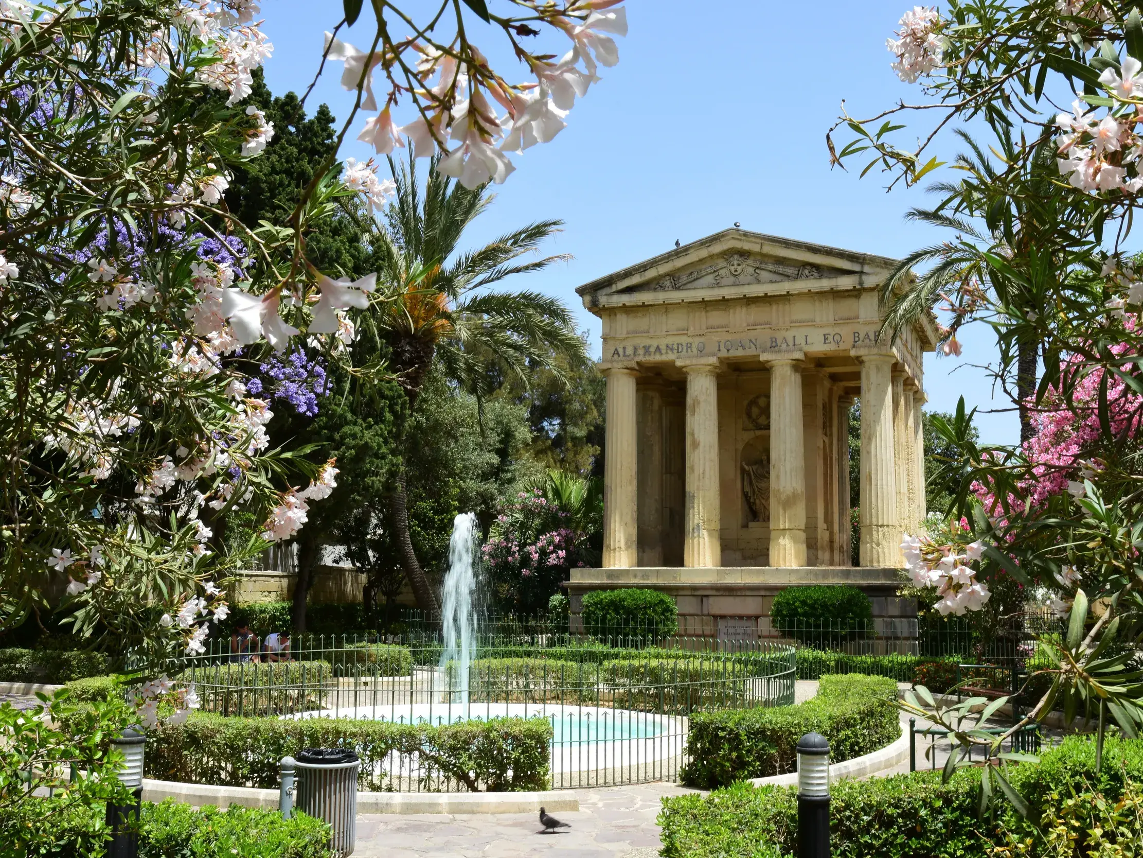 Klassizistisches Denkmal in den Lower Barakka Gardens in Valletta, eingerahmt von blühendem Oleander und mit einem Springbrunnen.