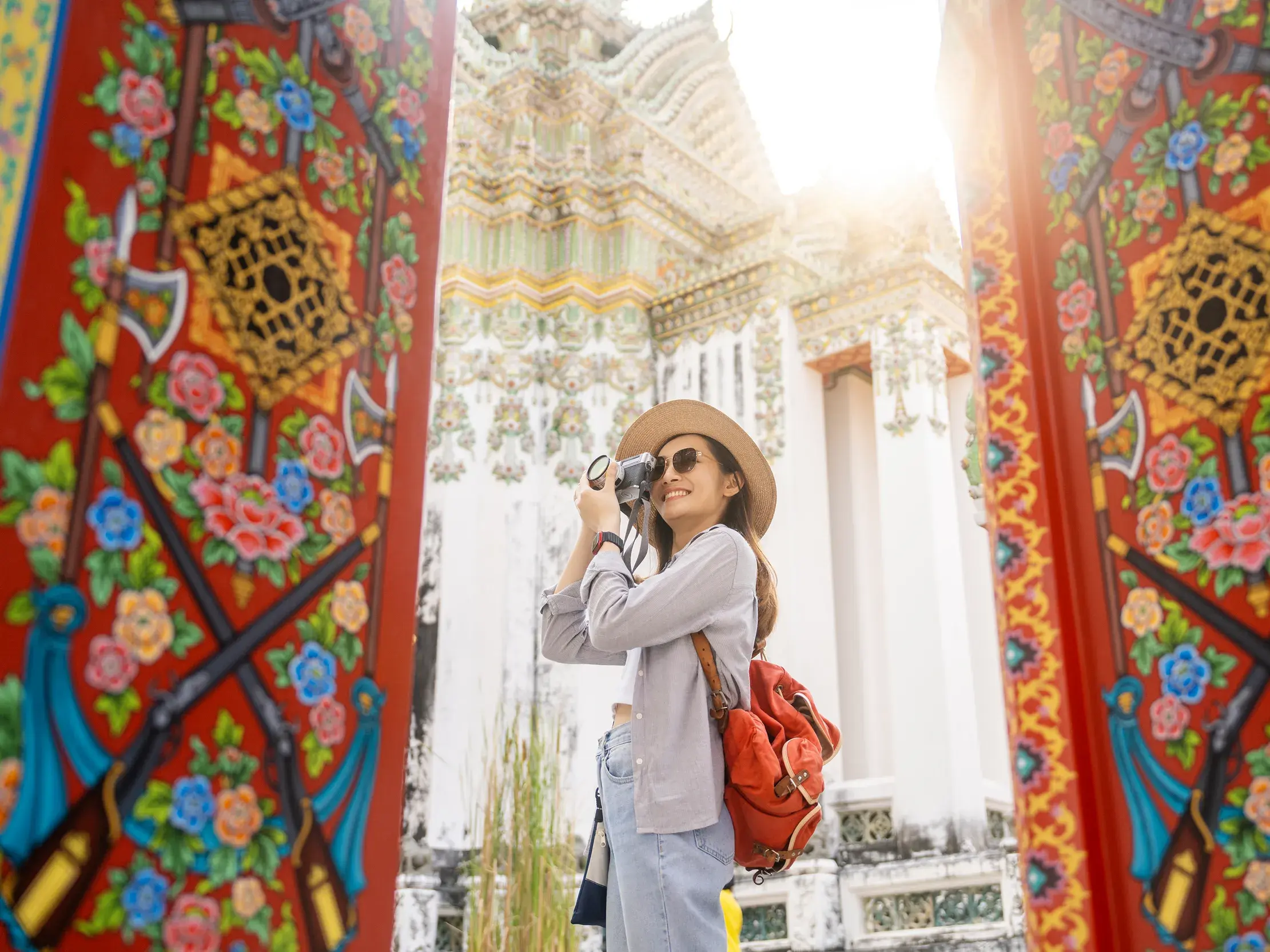 Alleinreisende mit Kamera zwischen reich verzierten Toren, die den sonnenbeschienenen Tempel Wat Arun in Bangkok einrahmen.