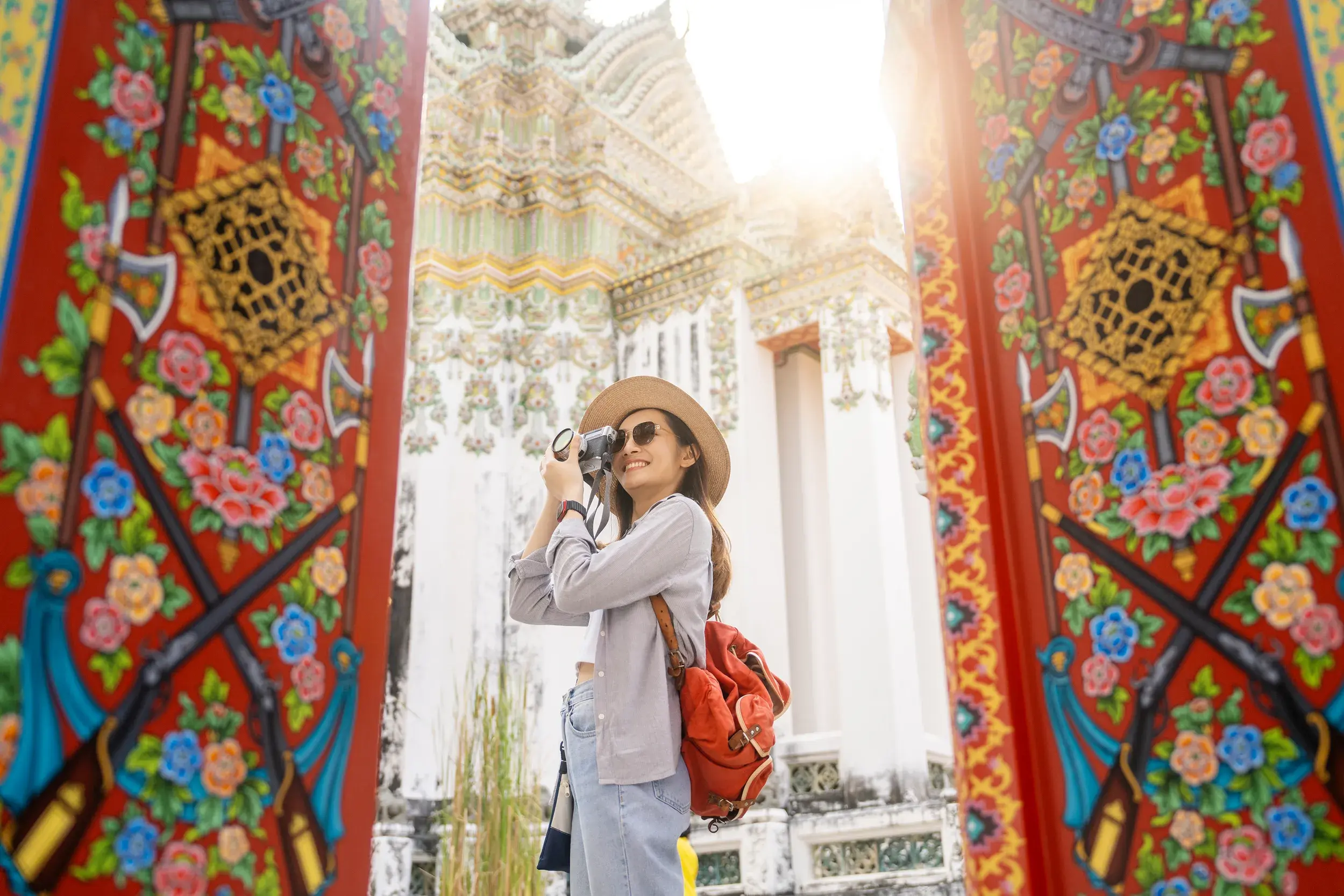 Alleinreisende mit Kamera zwischen reich verzierten Toren, die den sonnenbeschienenen Tempel Wat Arun in Bangkok einrahmen.
