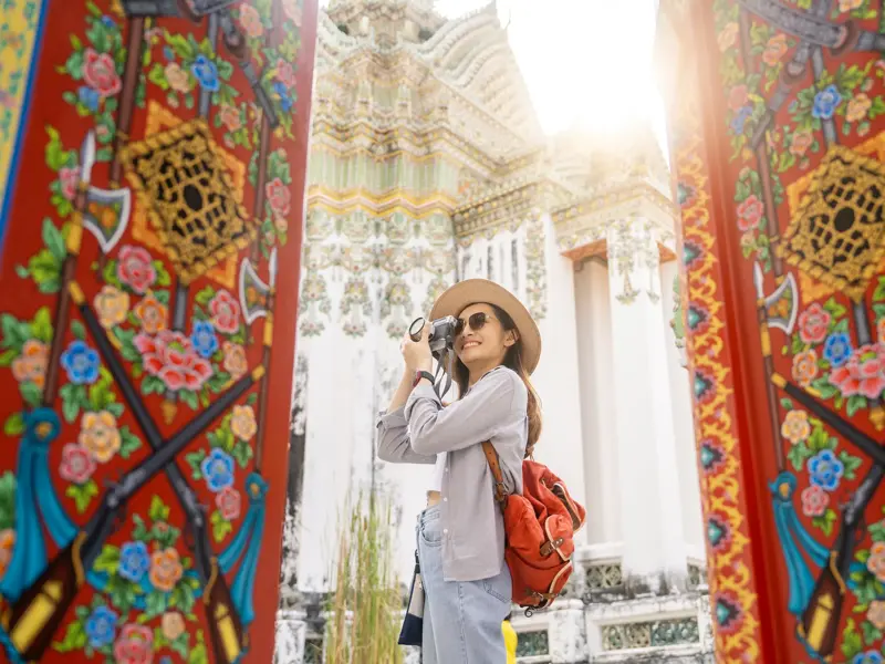 Alleinreisende mit Kamera zwischen reich verzierten Toren, die den sonnenbeschienenen Tempel Wat Arun in Bangkok einrahmen.