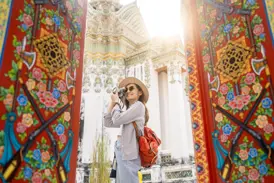 Alleinreisende mit Kamera zwischen reich verzierten Toren, die den sonnenbeschienenen Tempel Wat Arun in Bangkok einrahmen.