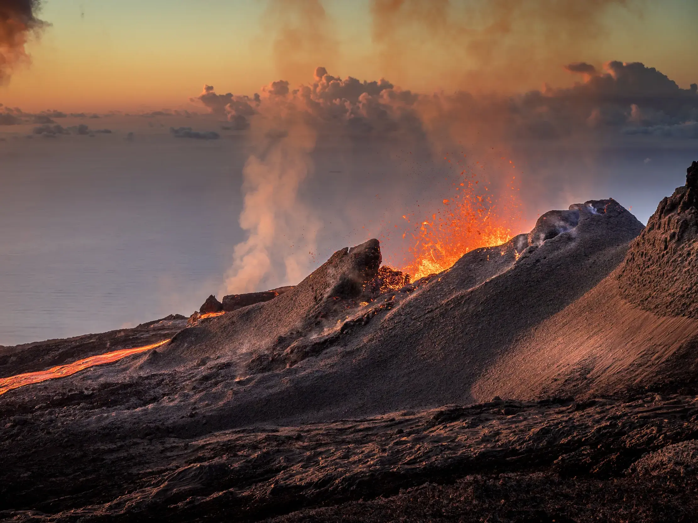 Ein Lavastrom ergießt sich vom Krater des Vulkans Piton de la Fournaise auf La Réunion, während Rauch in den Abendhimmel steigt.