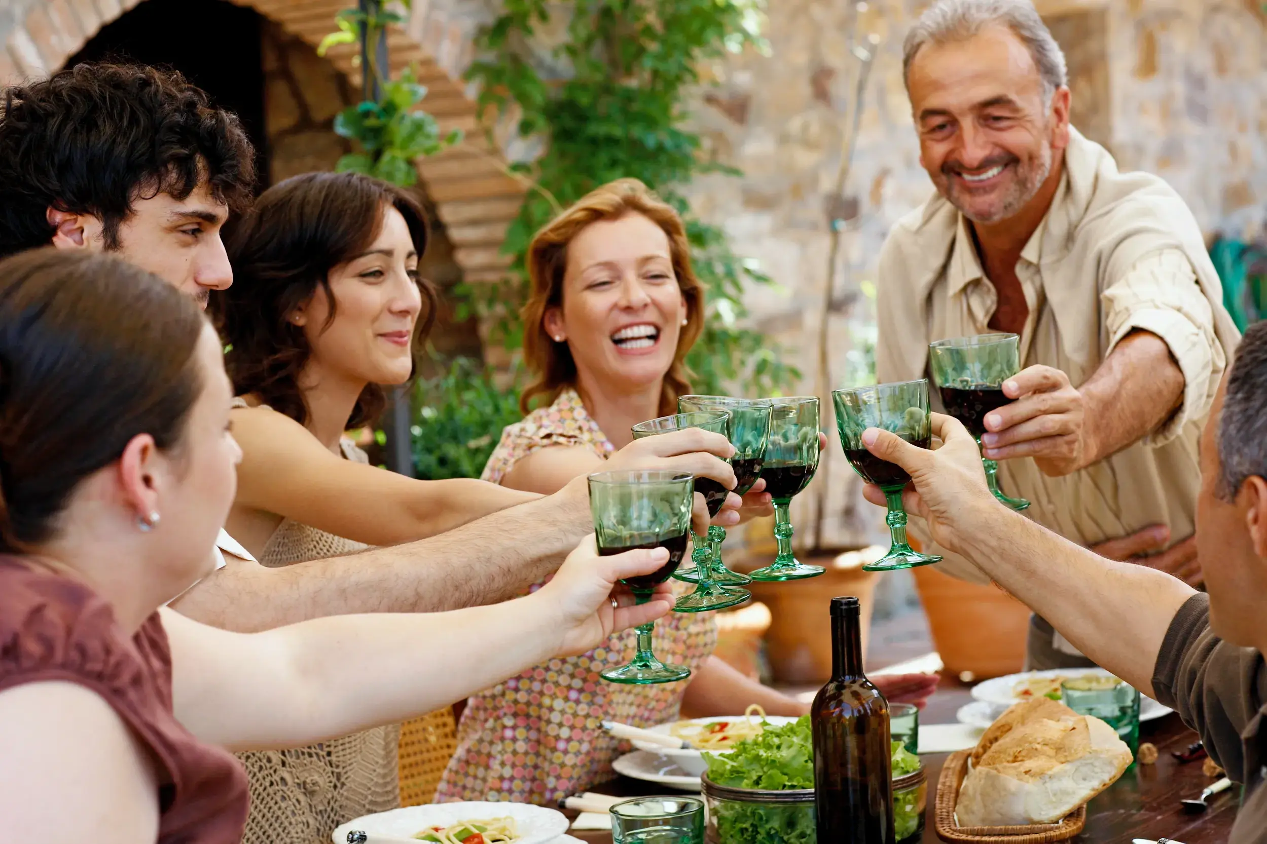 Eine fröhliche Reisegruppe stößt bei einem gemeinsamen Abendessen in einer rustikalen Masseria in Apulien mit Rotwein an.