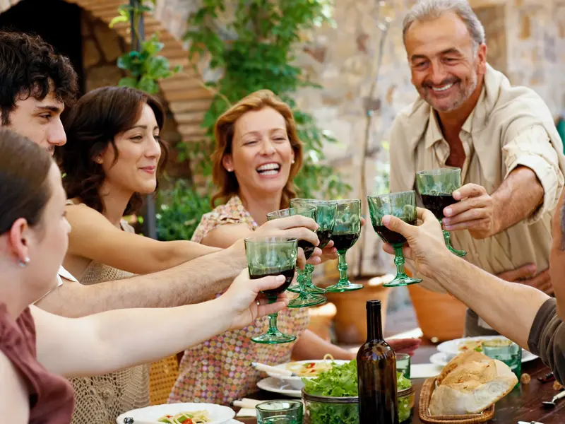 Eine fröhliche Reisegruppe stößt bei einem gemeinsamen Abendessen in einer rustikalen Masseria in Apulien mit Rotwein an.