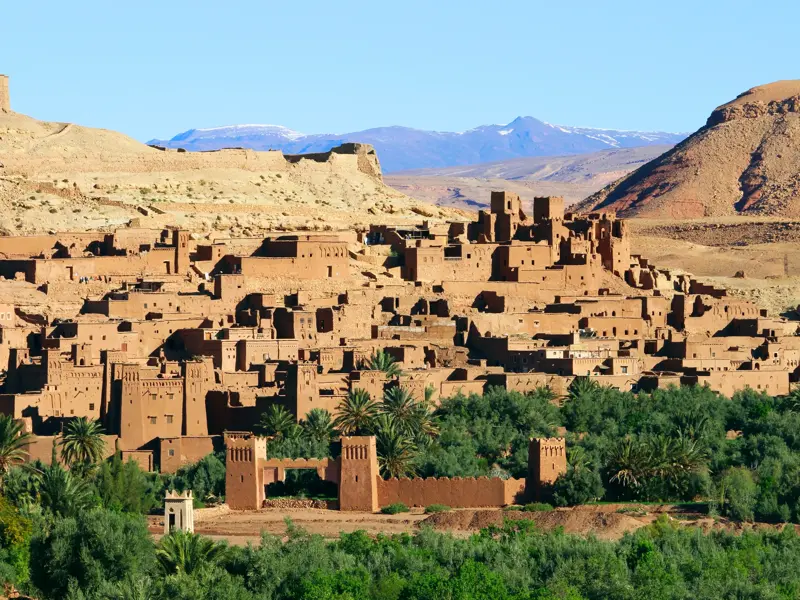 Die Lehmbausiedlung Ait Benhaddou in Marokko, eingebettet in eine grüne Oase vor dem schneebedeckten Hohen Atlas.
