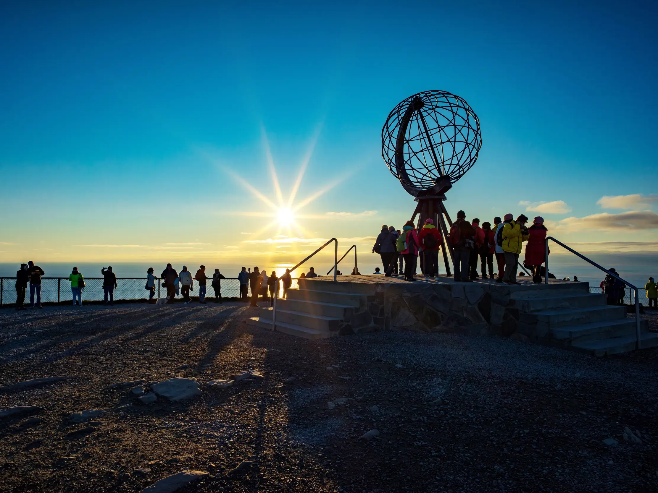 Eine Reisegruppe versammelt sich am Globus-Denkmal des Nordkap und blickt auf die strahlende Mitternachtssonne am Horizont.